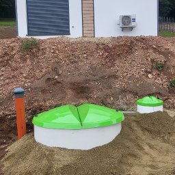 Z.P. H. MALWITZ Robert Malwic - Zainstalowana przydomowa oczyszczalnia ścieków z zielonymi pokrywami na tle budynku z klimatyzacją i roletą antywłamaniową. Widoczne rury i zasypka piaskowa.