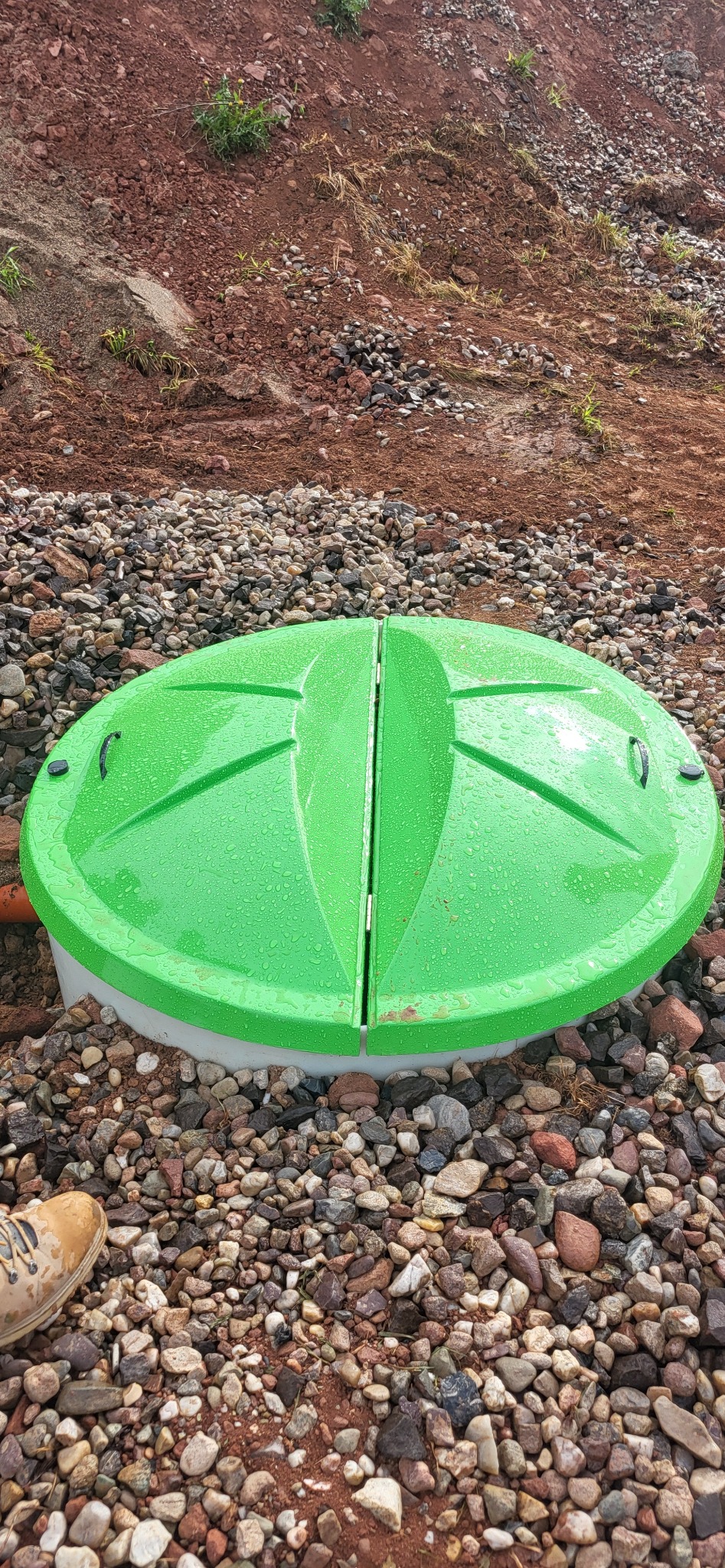 Zielona, dwuczęściowa pokrywa przydomowej oczyszczalni ścieków na żwirowym podłożu, z fragmentem rury odpływowej i butem w rogu kadru. Widoczne krople wody na pokrywie.