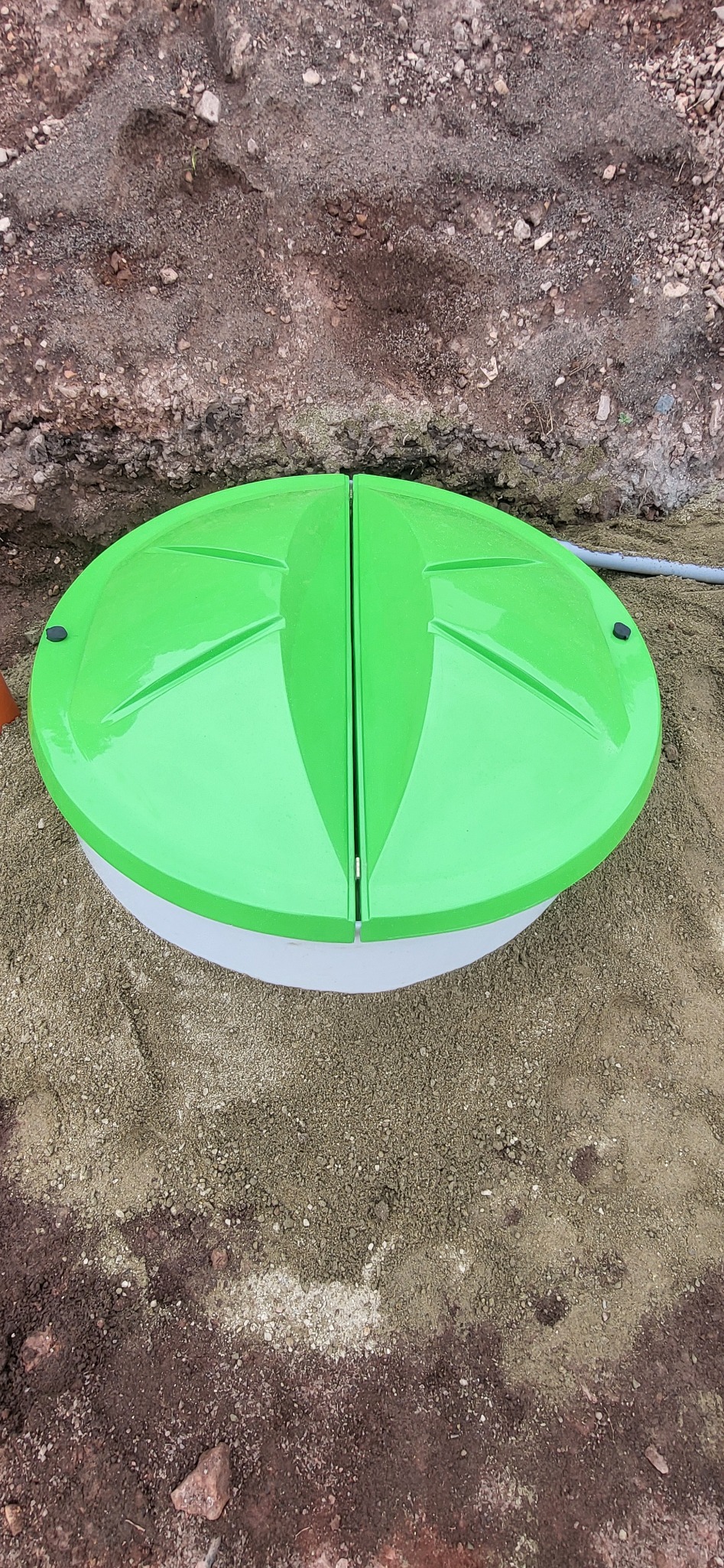 Zielona pokrywa przydomowej oczyszczalni ścieków, widoczna z góry, otoczona piaskiem i ziemią. Dwu-częściowa pokrywa z wyraźnym podziałem na środku.