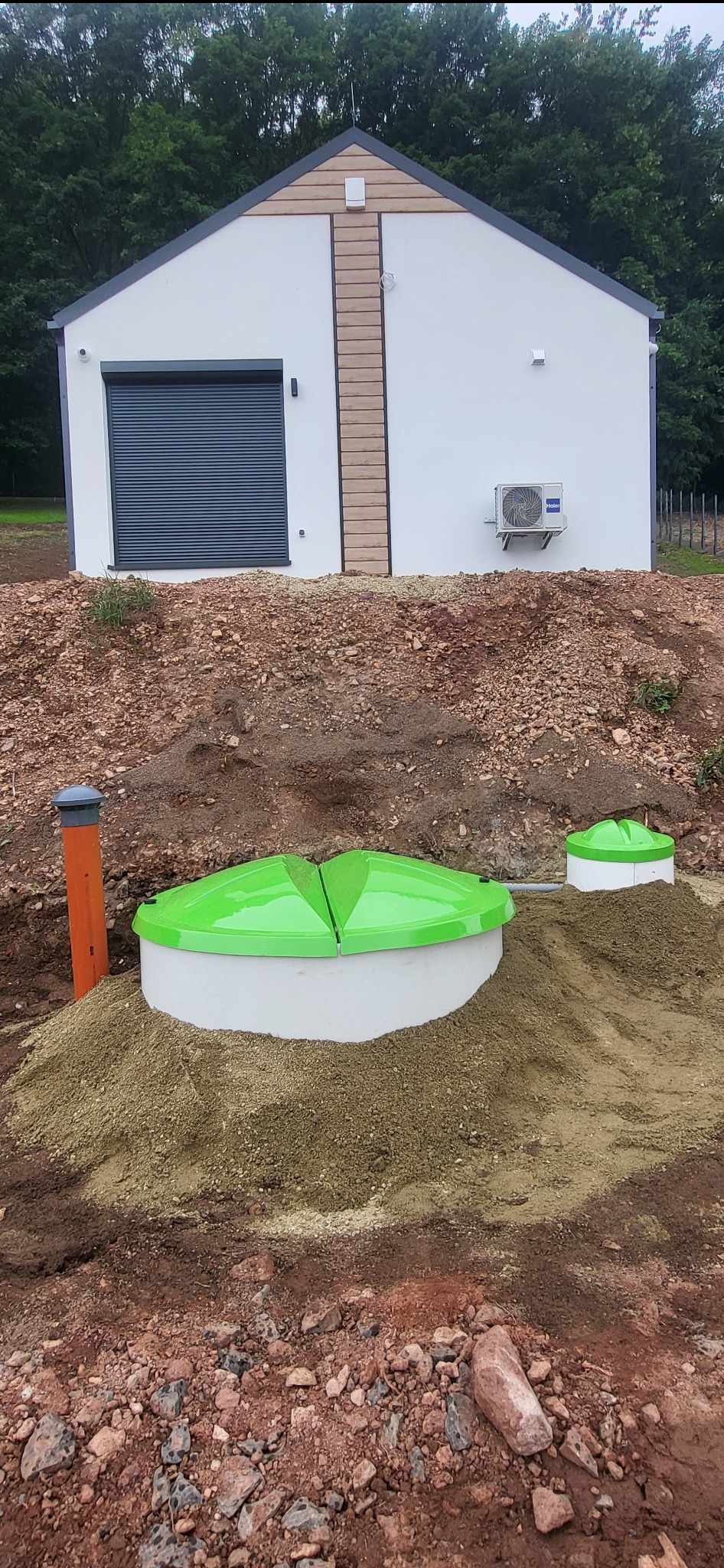 Zainstalowana przydomowa oczyszczalnia ścieków z zielonymi pokrywami na tle budynku z klimatyzacją i roletą antywłamaniową. Widoczne rury i zasypka piaskowa.