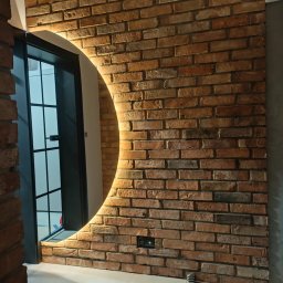 Jakub Porządnicki Porządny Remont - Ceglana ściana z okrągłym lustrem podświetlanym LED, czarne gniazdko elektryczne, fragment drzwi z czarną ramą. Ciepłe oświetlenie podkreśla fakturę cegieł.