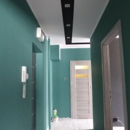 Jakub Porządnicki Porządny Remont - Pomalowany na zielono korytarz z szarą podłogą imitującą drewno. Na suficie czarny pas z oświetleniem punktowym. Widoczne drzwi i elementy instalacji.