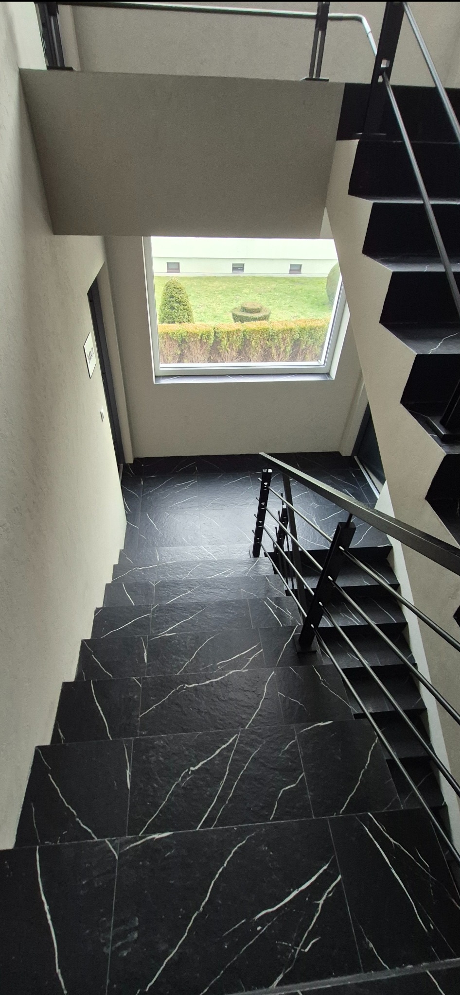 Schody wyłożone czarnym gresem z białym żyłkowaniem, widok z góry. Metalowa barierka. W tle okno z widokiem na zieleń. Minimalistyczny design.