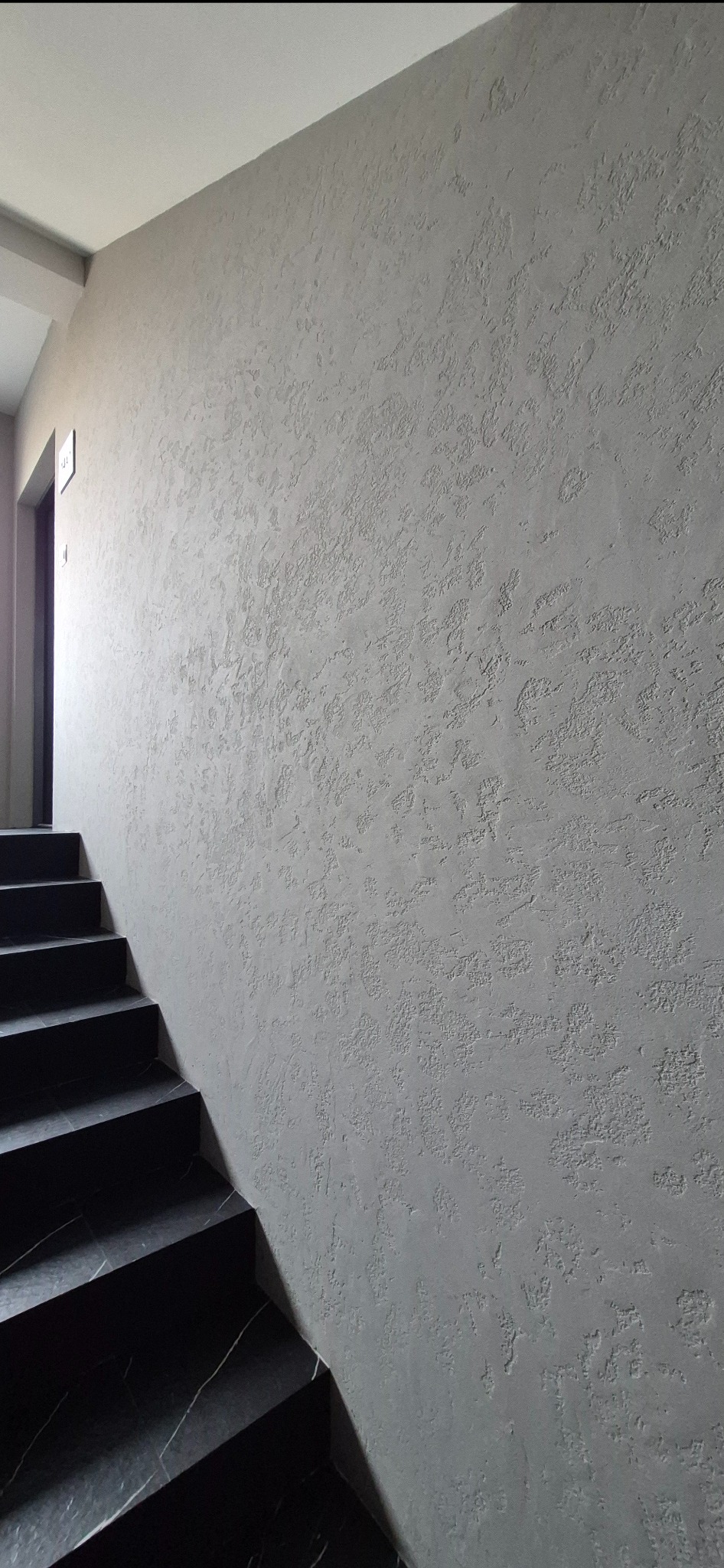 Szara ściana z dekoracyjną fakturą w korytarzu, obok schody z czarnego marmuru. Widoczne wejście do pomieszczenia. Minimalistyczny design.