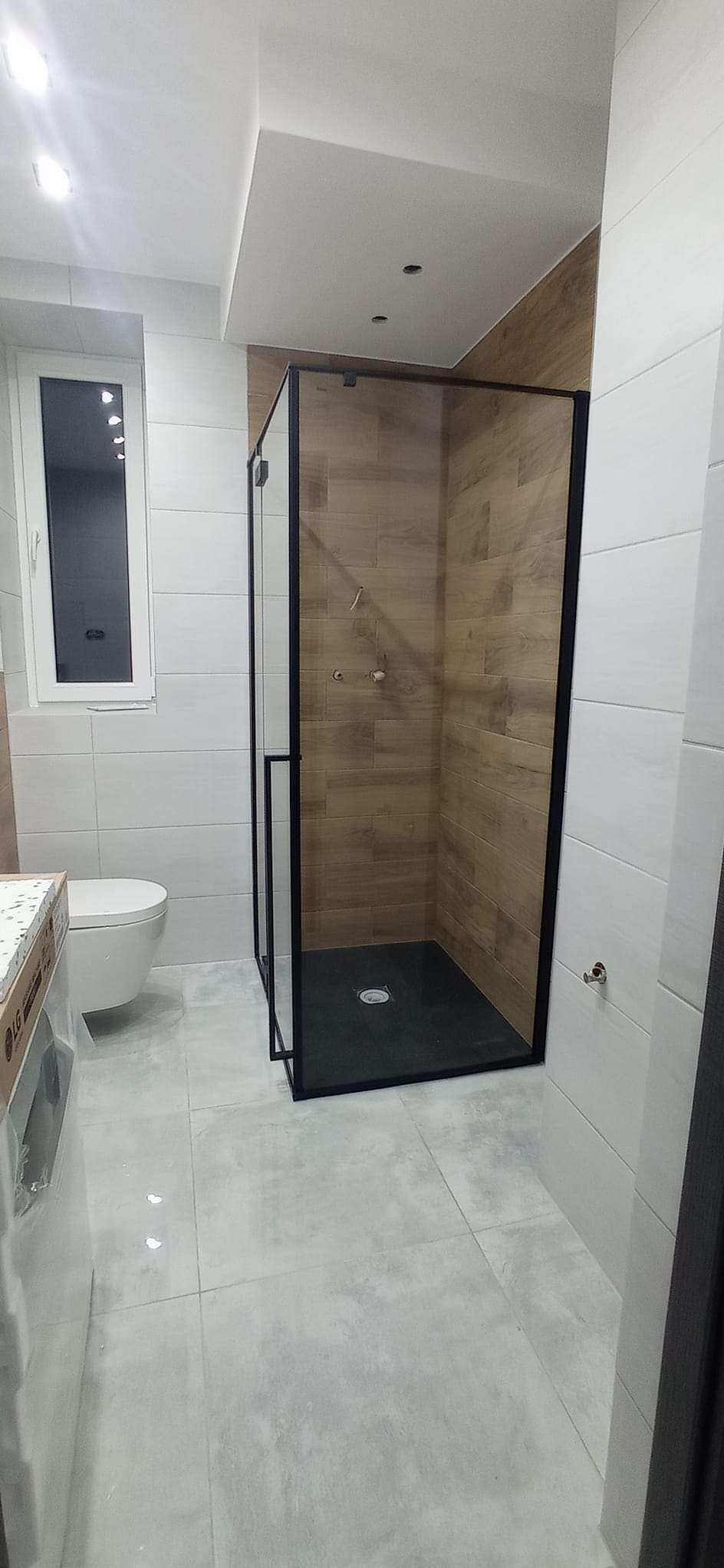 Nowoczesna łazienka po remoncie: kabina prysznicowa z czarną ramą, drewnopodobne płytki w strefie prysznica, białe płytki na ścianach, szara podłoga i toaleta.