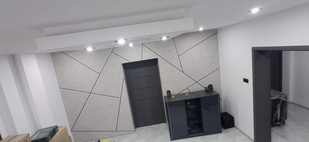 Wykończona ściana z geometrycznym wzorem z płytek, szara szafka RTV i drzwi w nowoczesnym wnętrzu. Oświetlenie sufitowe punktowe.