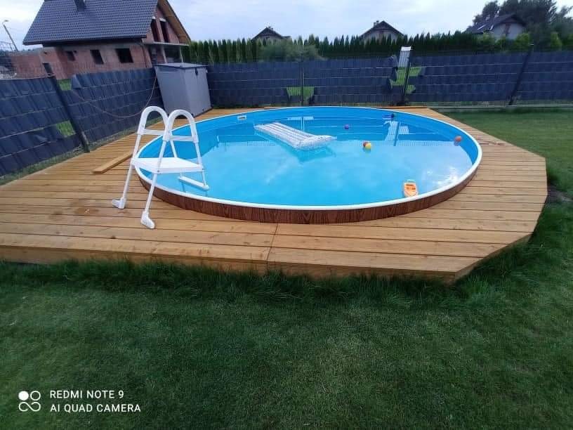Okrągły basen ogrodowy z drewnianym tarasem, drabinką i akcesoriami do pływania na trawniku. W tle dom i ogrodzenie.
