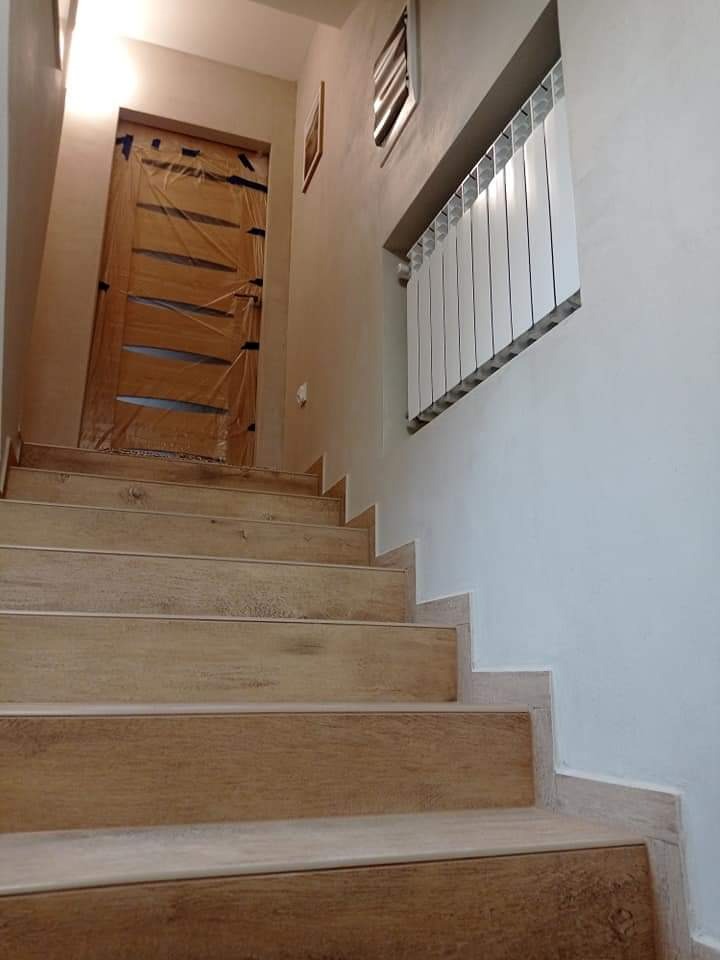 Wykończone schody z drewnopodobnymi płytkami, jasne ściany, grzejnik pod oknem. Drzwi zabezpieczone folią ochronną. Ujęcie od dołu, widok na górę schodów.