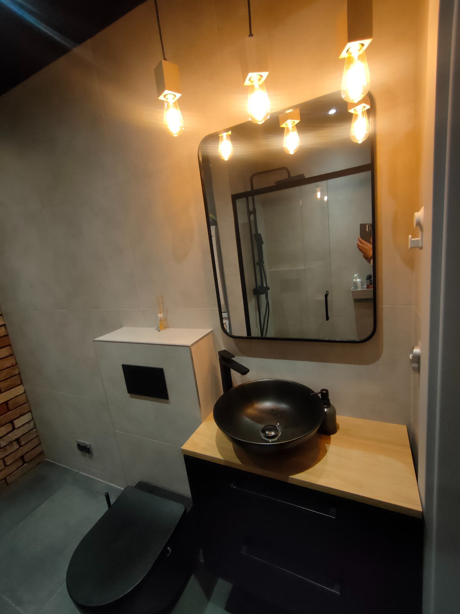 Nowoczesna łazienka z czarną umywalką na drewnianym blacie, minimalistycznym oświetleniem i lustrem odbijającym kabinę prysznicową. Szare płytki i ceglana ściana.