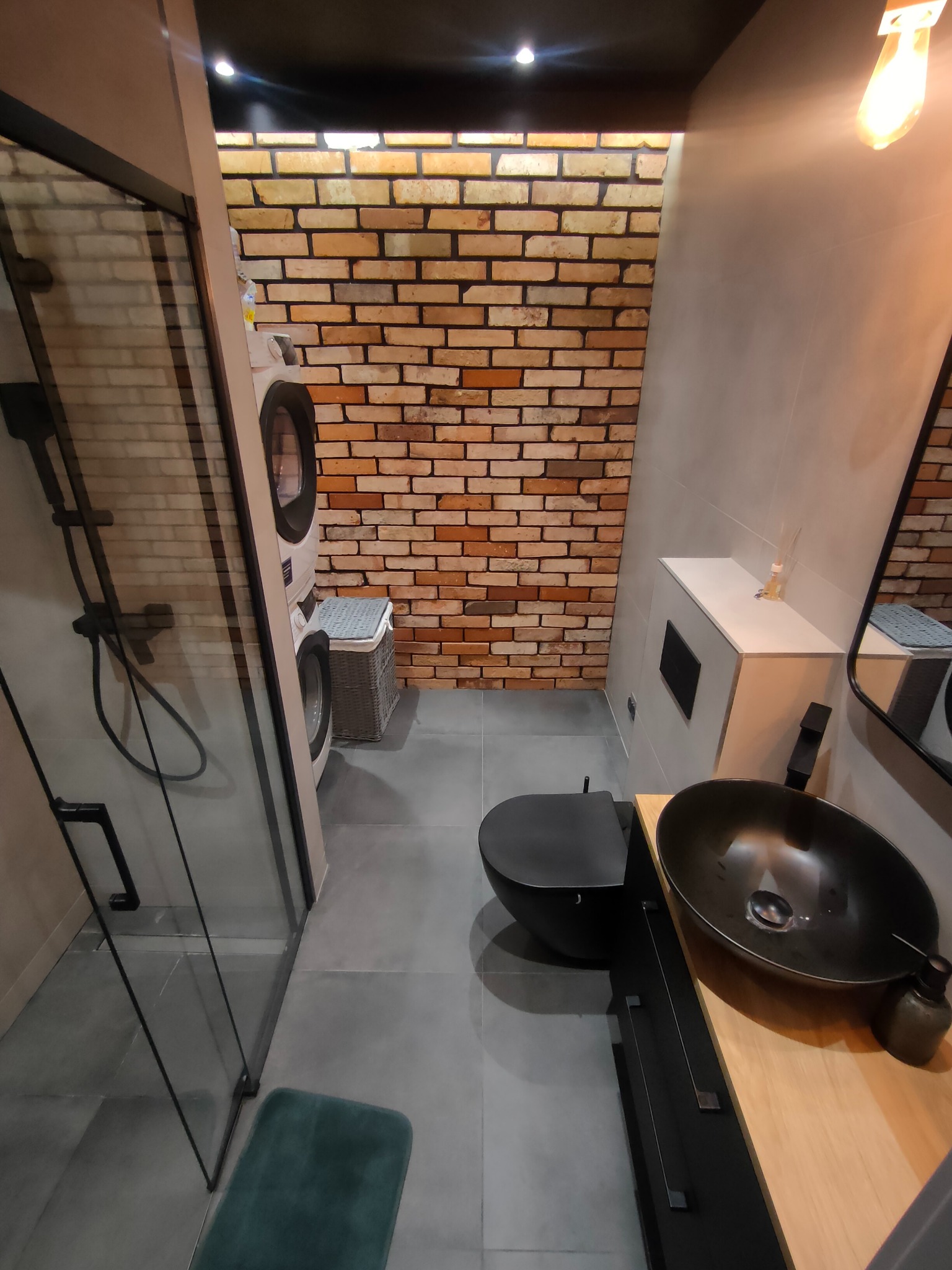 Nowoczesna łazienka z kabiną prysznicową typu walk-in, umywalką nablatową i ścianą z cegły. Widoczna pralka i kosz na pranie. Oświetlenie punktowe.
