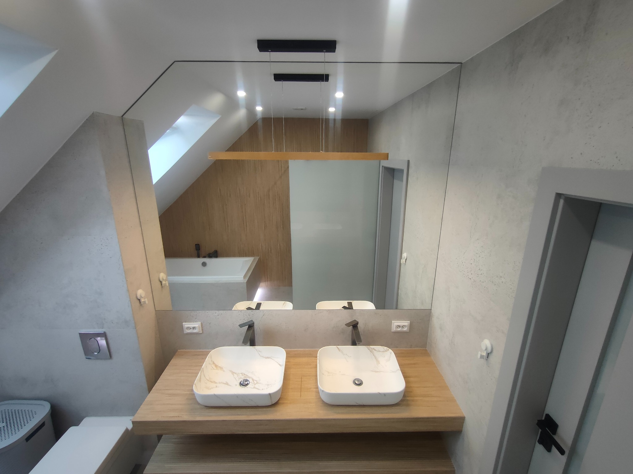 Nowoczesna łazienka na poddaszu z dwoma umywalkami nablatowymi, dużym lustrem i oświetleniem LED. Widoczna wanna w tle. Ściany wykończone w szarym betonie.
