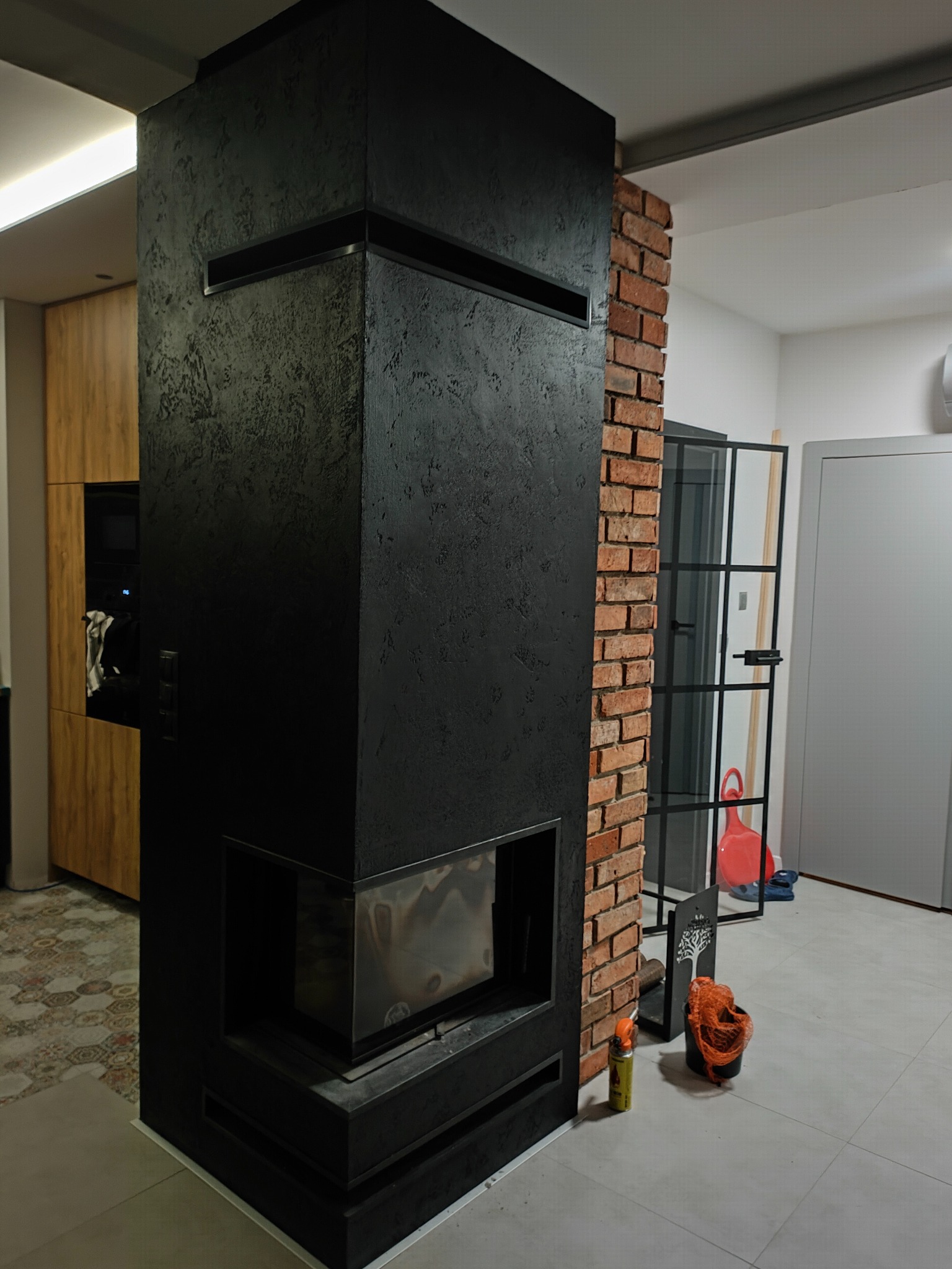 Nowoczesny kominek z czarną, teksturowaną obudową i ceglanym wykończeniem ściany. Widoczny fragment kuchni z drewnianymi szafkami i wzorzystą podłogą.