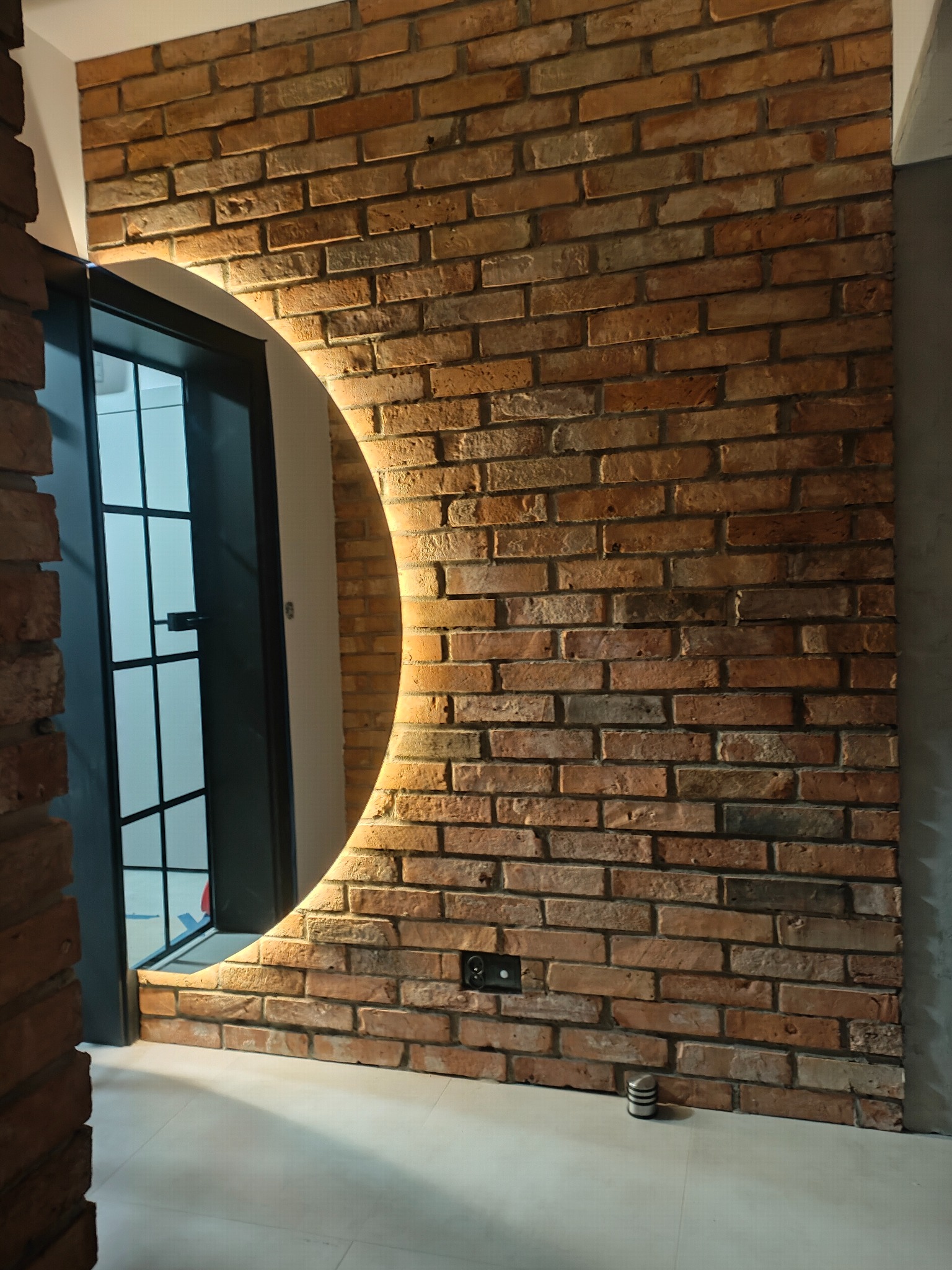 Ceglana ściana z okrągłym lustrem podświetlanym LED, czarne gniazdko elektryczne, fragment drzwi z czarną ramą. Ciepłe oświetlenie podkreśla fakturę cegieł.