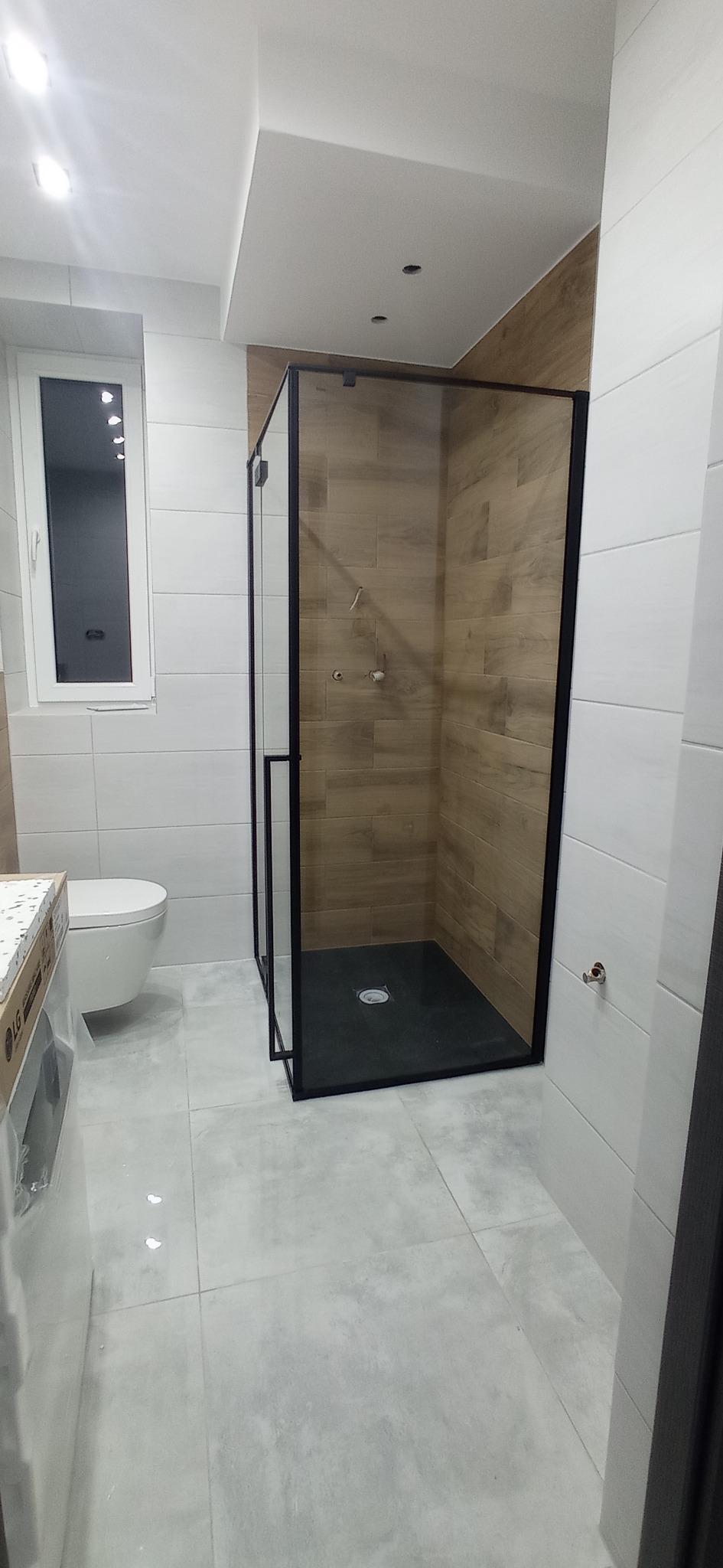 Nowoczesna łazienka z kabiną prysznicową typu walk-in z czarnymi profilami, wykończona płytkami imitującymi drewno i szarą podłogą. Widoczna toaleta i okno.