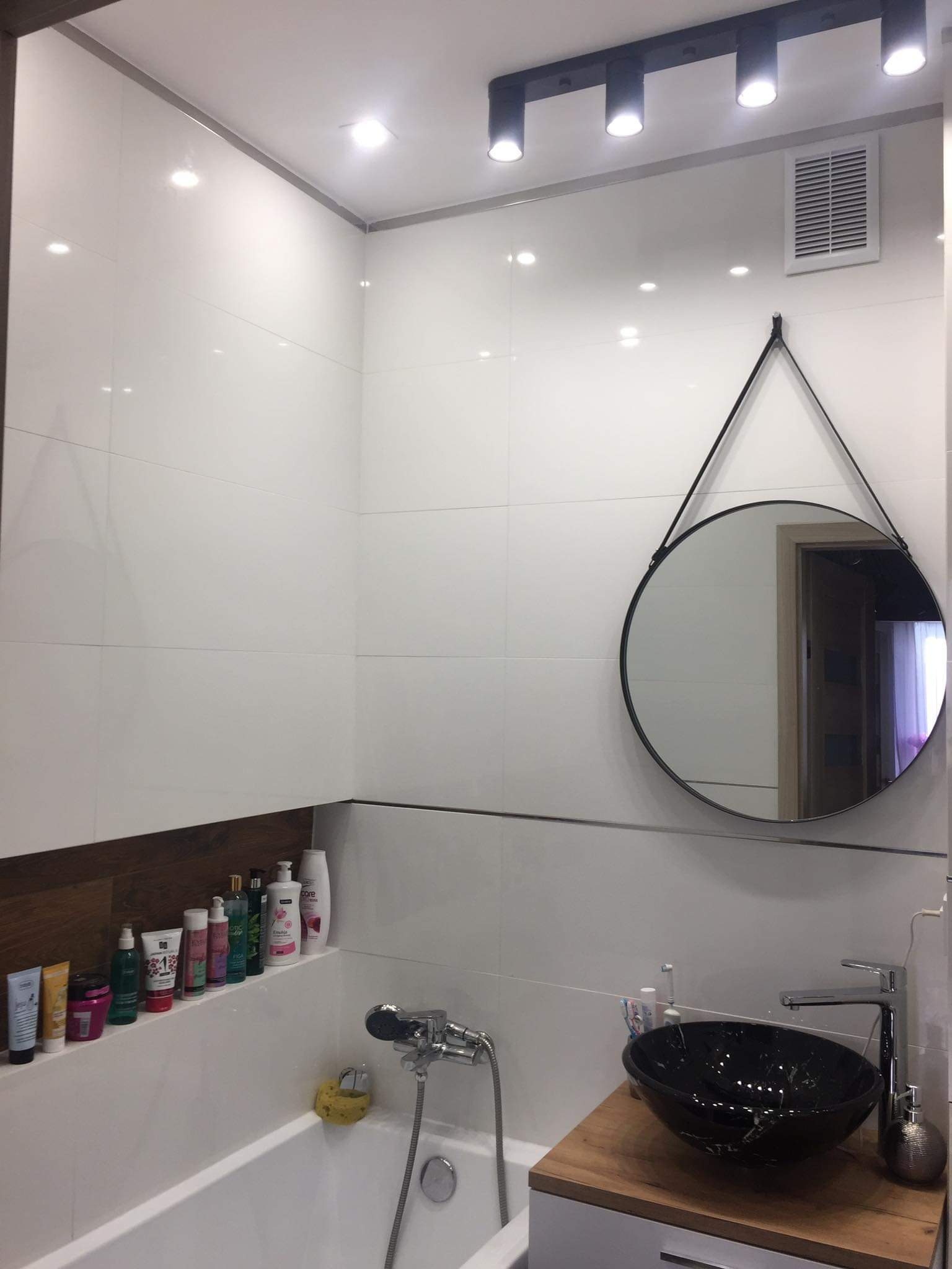 Biała łazienka z czarną umywalką na drewnianym blacie, okrągłe lustro na ścianie z białych płytek, oświetlenie punktowe i wanna z kosmetykami.