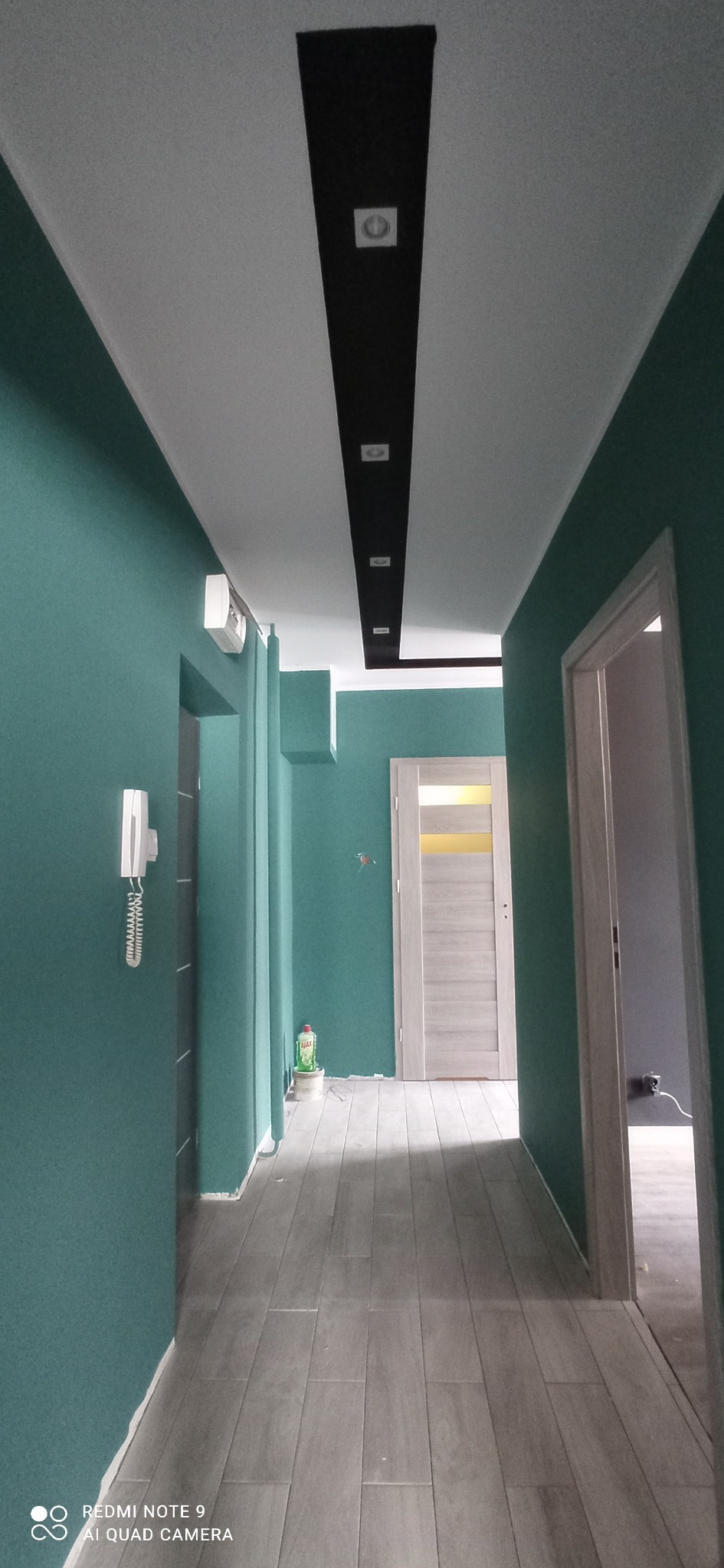 Pomalowany na zielono korytarz z szarą podłogą imitującą drewno. Na suficie czarny pas z oświetleniem punktowym. Widoczne drzwi i elementy instalacji.