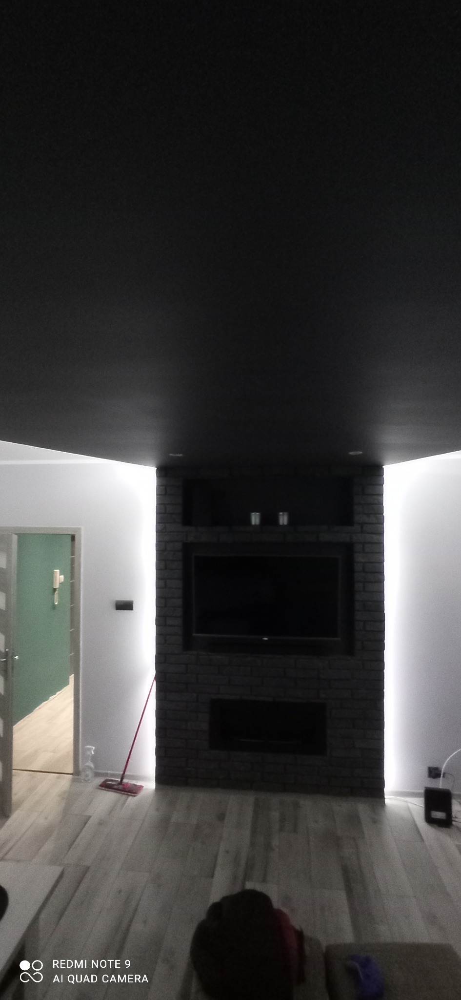 Ciemna ściana z cegły z wnęką na telewizor i oświetleniem LED, na tle jasnych ścian i podłogi. Widoczny fragment sufitu i drzwi do innego pomieszczenia.