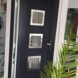 Katarzyna Zhadan - Ciemnoszare drzwi zewnętrzne z trzema kwadratowymi przeszkleniami w srebrnych ramkach, osadzone w białej futrynie, widoczne przez liście rośliny.