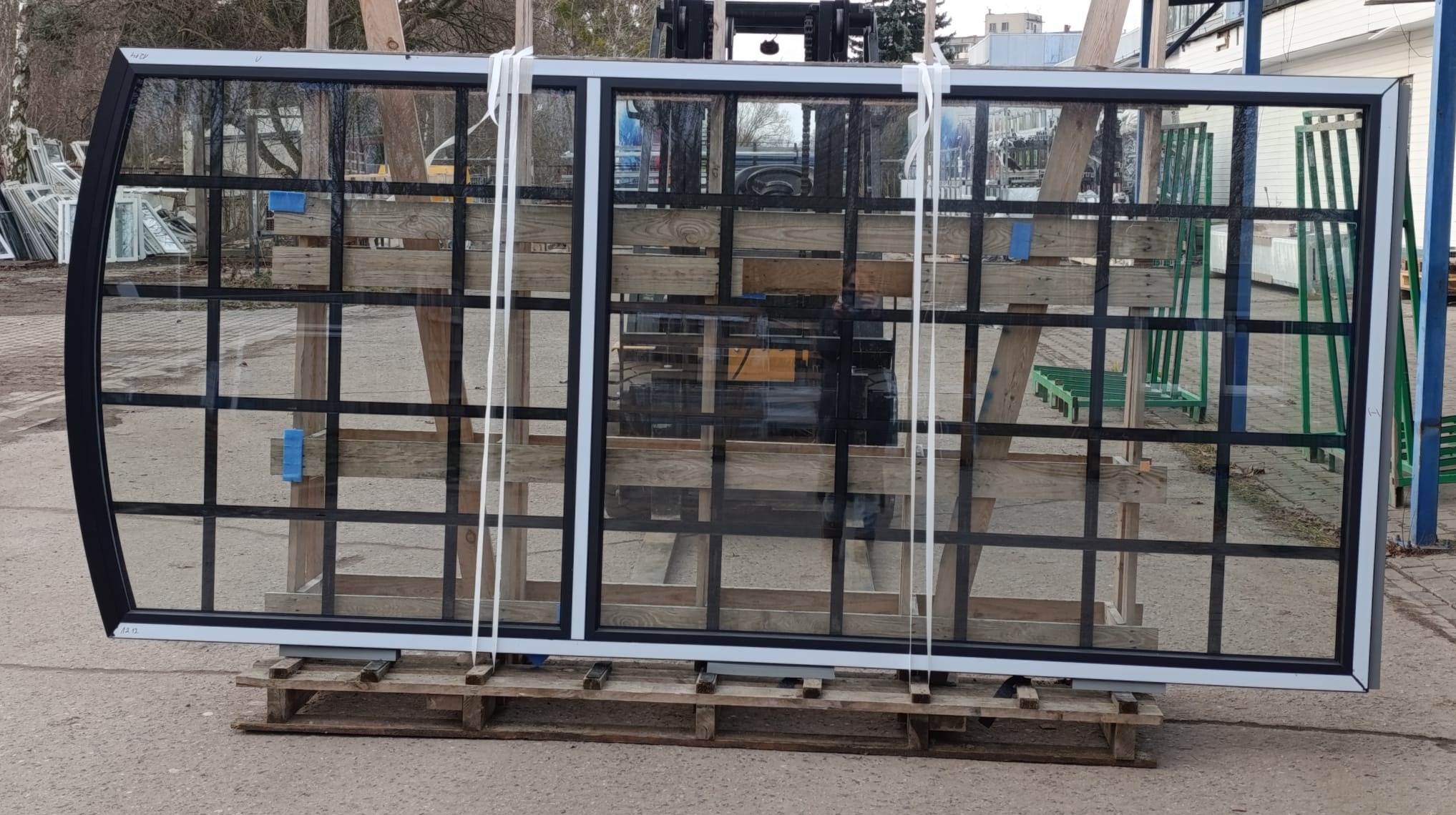 Nowe okno z czarną ramą i szprosami, zapakowane na palecie i zabezpieczone taśmami, czeka na montaż. W tle widać wózek widłowy i inne okna.