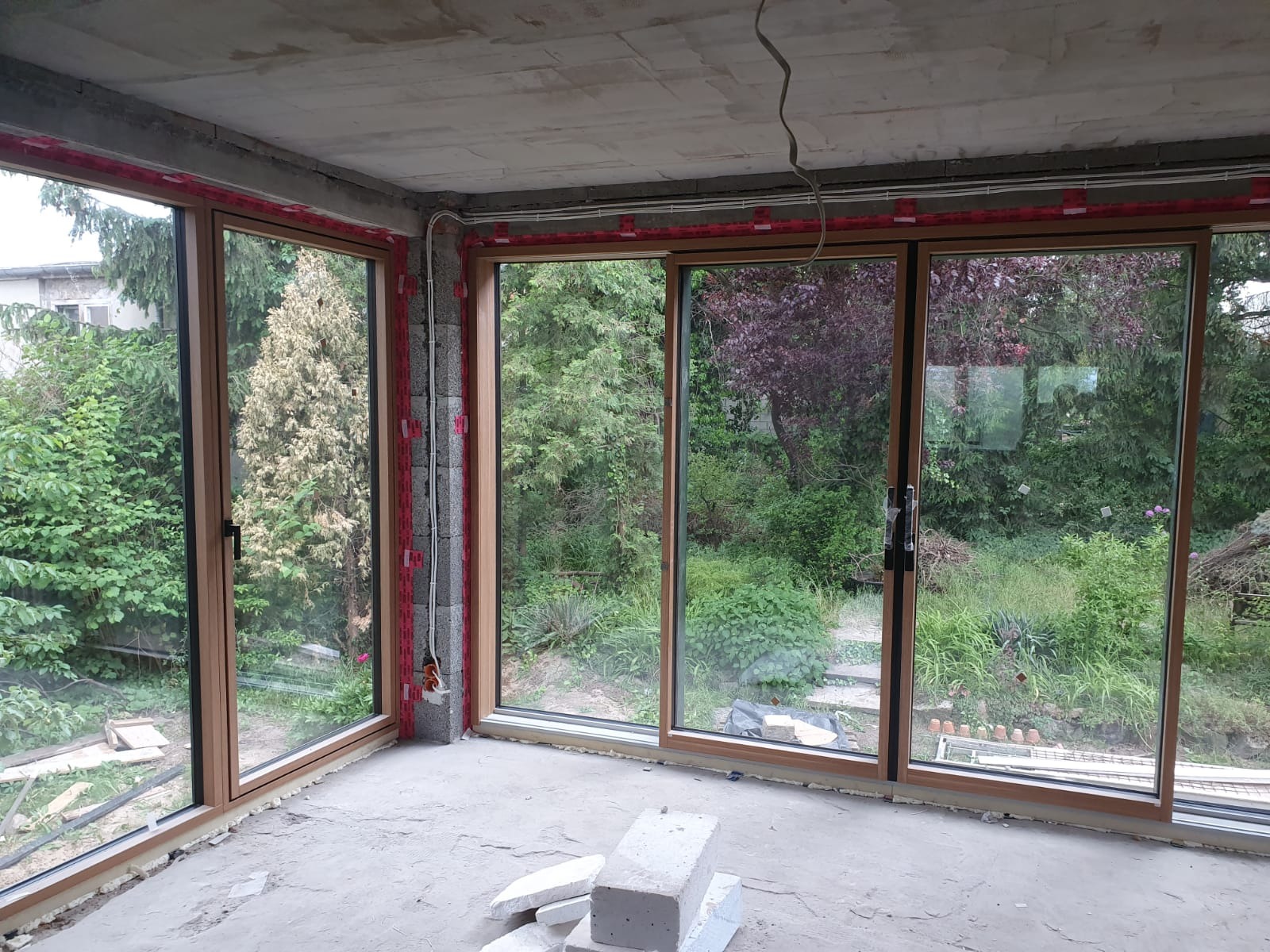 Wnętrze w stanie surowym z dużymi, przesuwnymi oknami z brązowymi ramami, wychodzącymi na bujny ogród. Widoczne elementy montażowe i instalacje elektryczne.