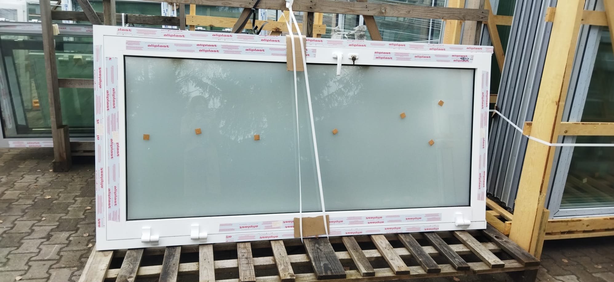 Białe okno PCV marki Aliplast, zabezpieczone piankowymi podkładkami, leży na drewnianej palecie. W tle widoczne inne okna.