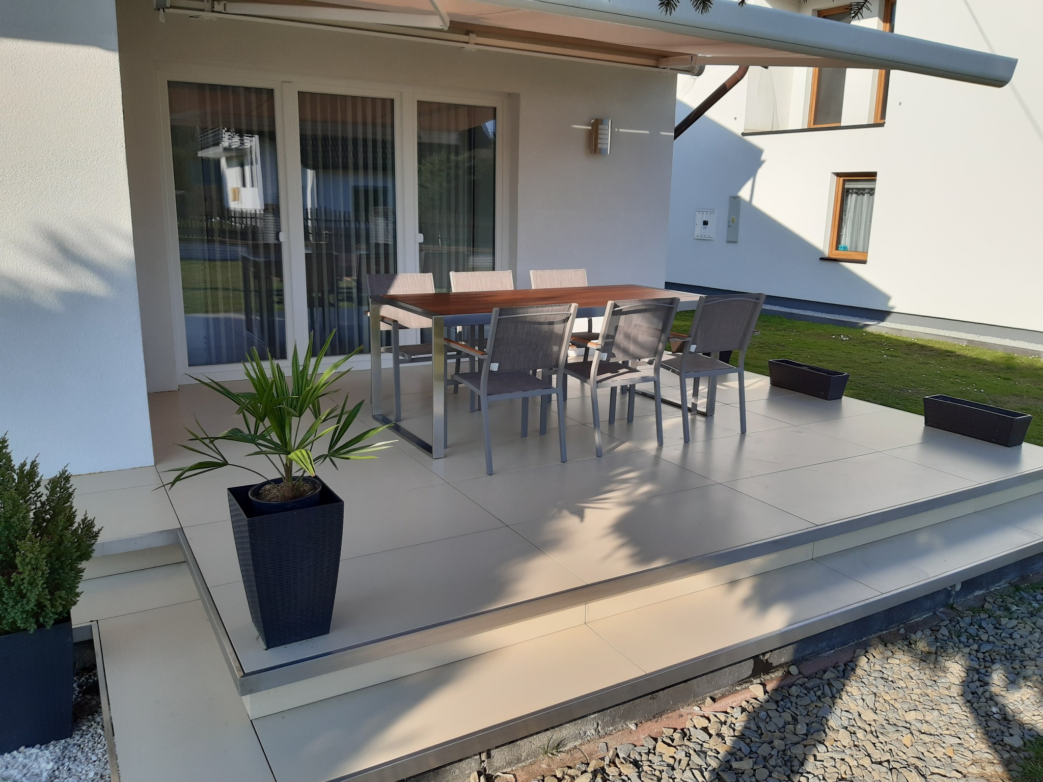 Nowoczesny taras z jasnymi płytkami, meblami ogrodowymi i roślinami w donicach. Widoczne schody tarasowe i markiza. Minimalistyczny design.