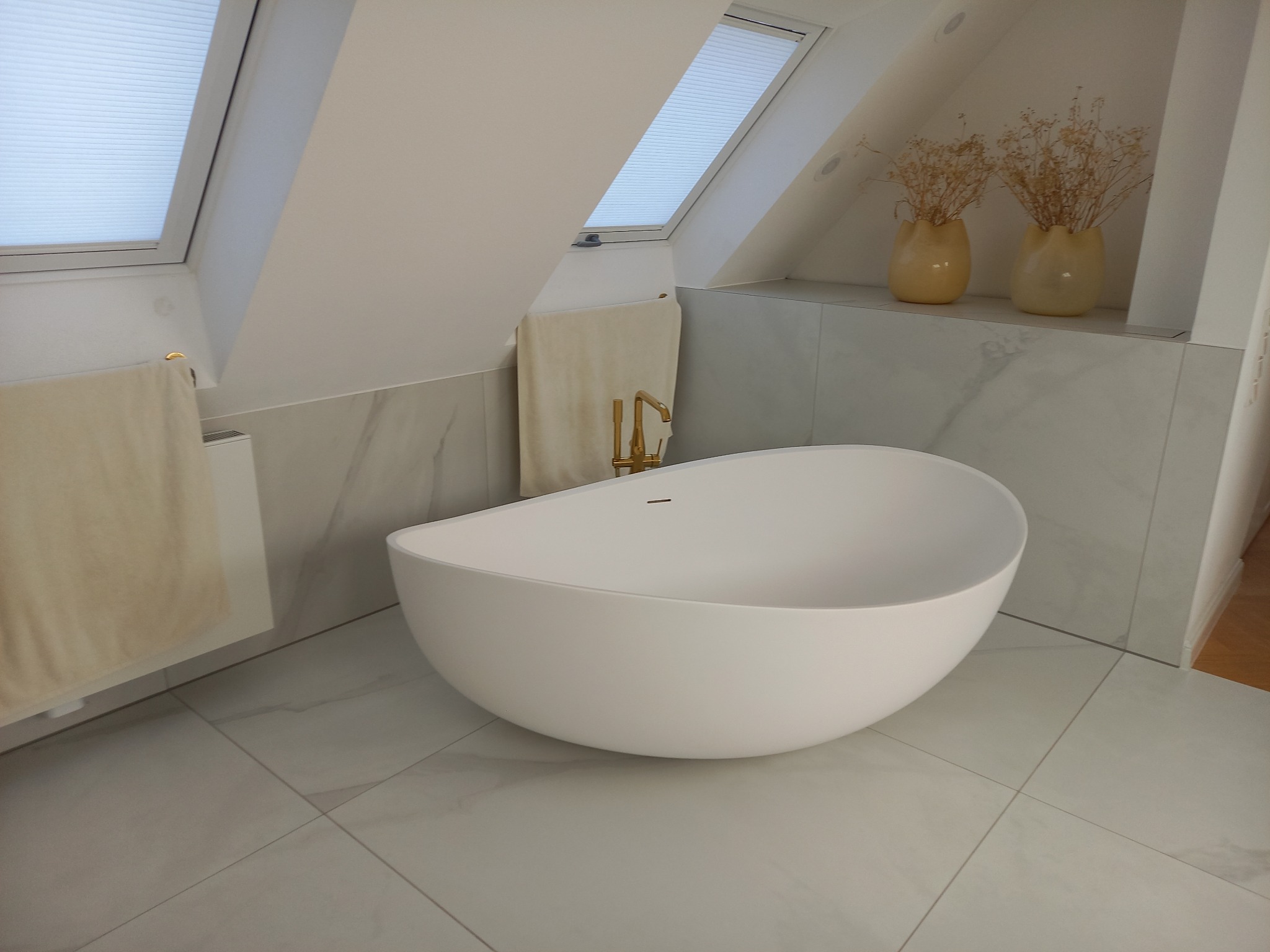 Nowoczesna, biała wanna wolnostojąca w jasnej łazience na poddaszu z oknami dachowymi i marmurowymi płytkami. Złota bateria, beżowe ręczniki i dekoracyjne wazony.