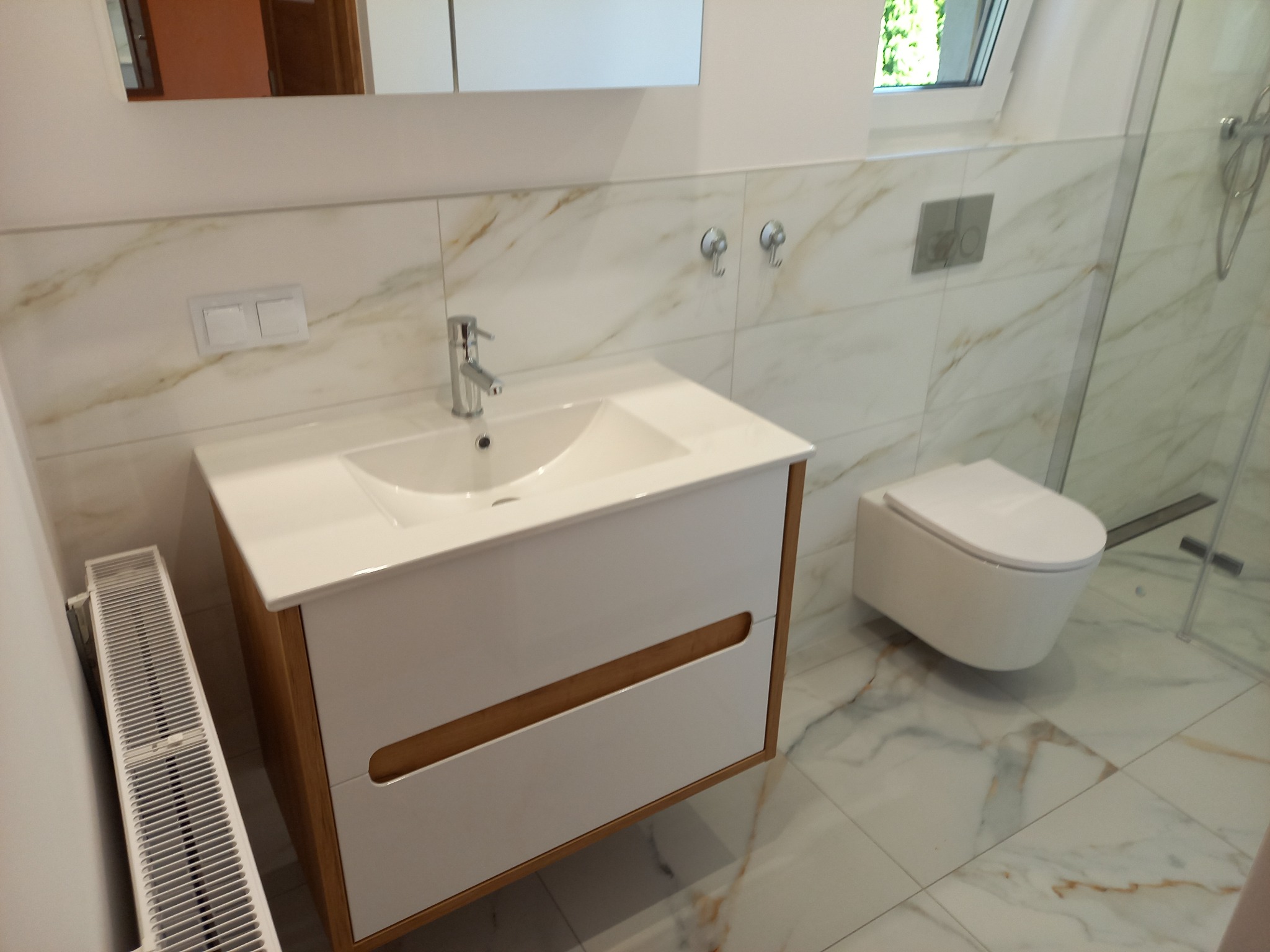 Nowoczesna łazienka z marmurowymi płytkami, wiszącą toaletą i szafką pod umywalkę z drewnianymi akcentami. Widoczny grzejnik i kabina prysznicowa.