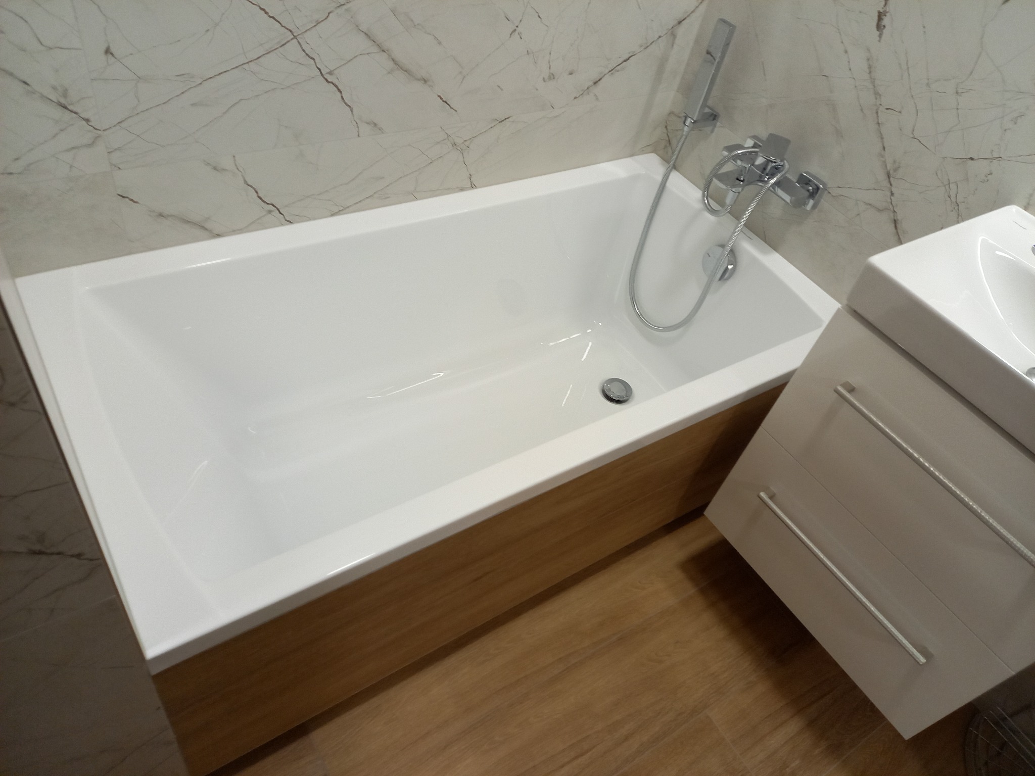 Biała wanna z obudową imitującą drewno, chromowana bateria z prysznicem, umywalka z szafką w łazience wykończonej marmurowymi płytkami. Nowoczesny design.