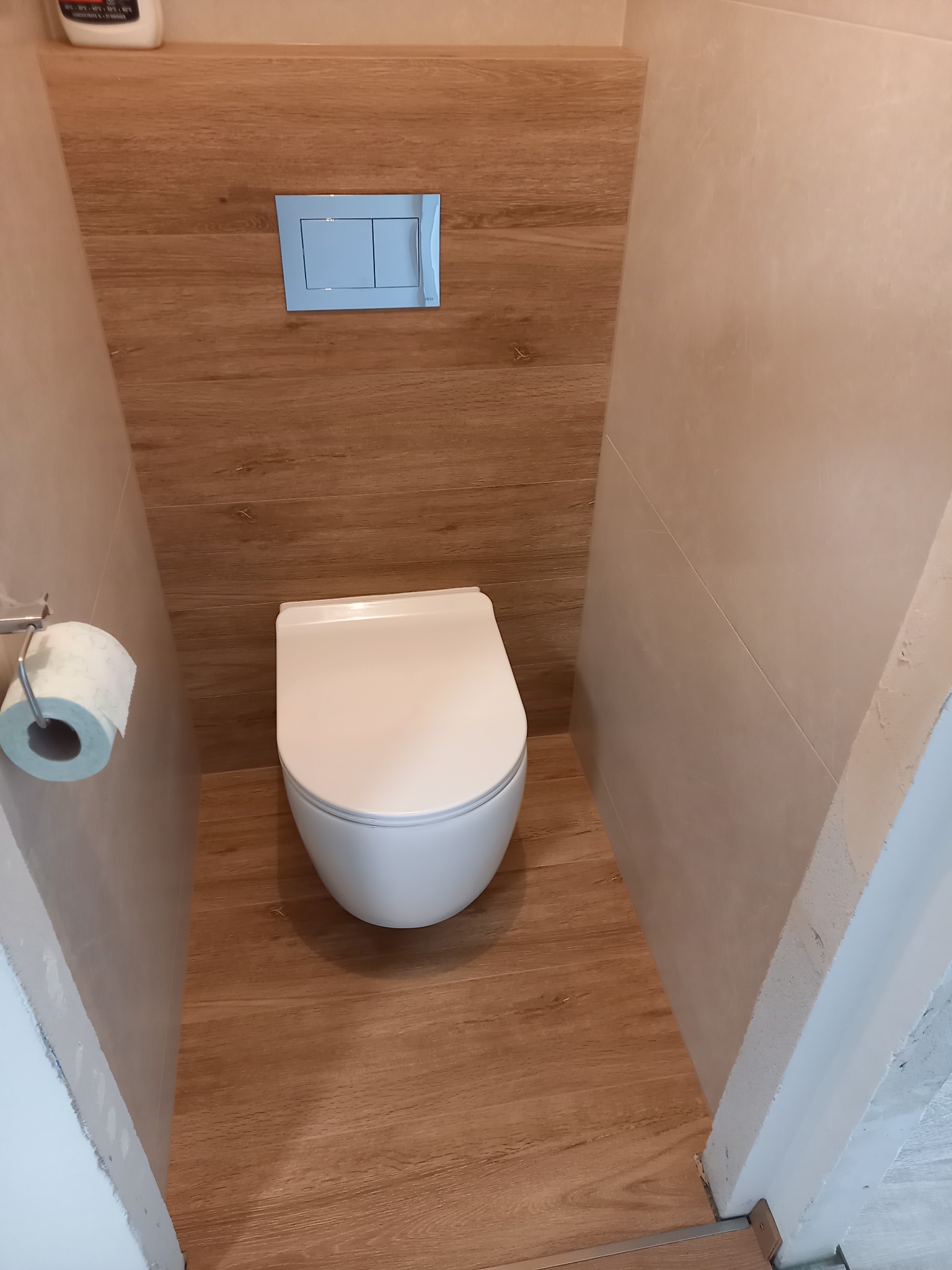 Kompaktowa toaleta w zabudowie z drewnopodobnymi płytkami, wisząca miska WC i niebieski przycisk spłukujący. Widoczny fragment ściany z surowym wykończeniem.