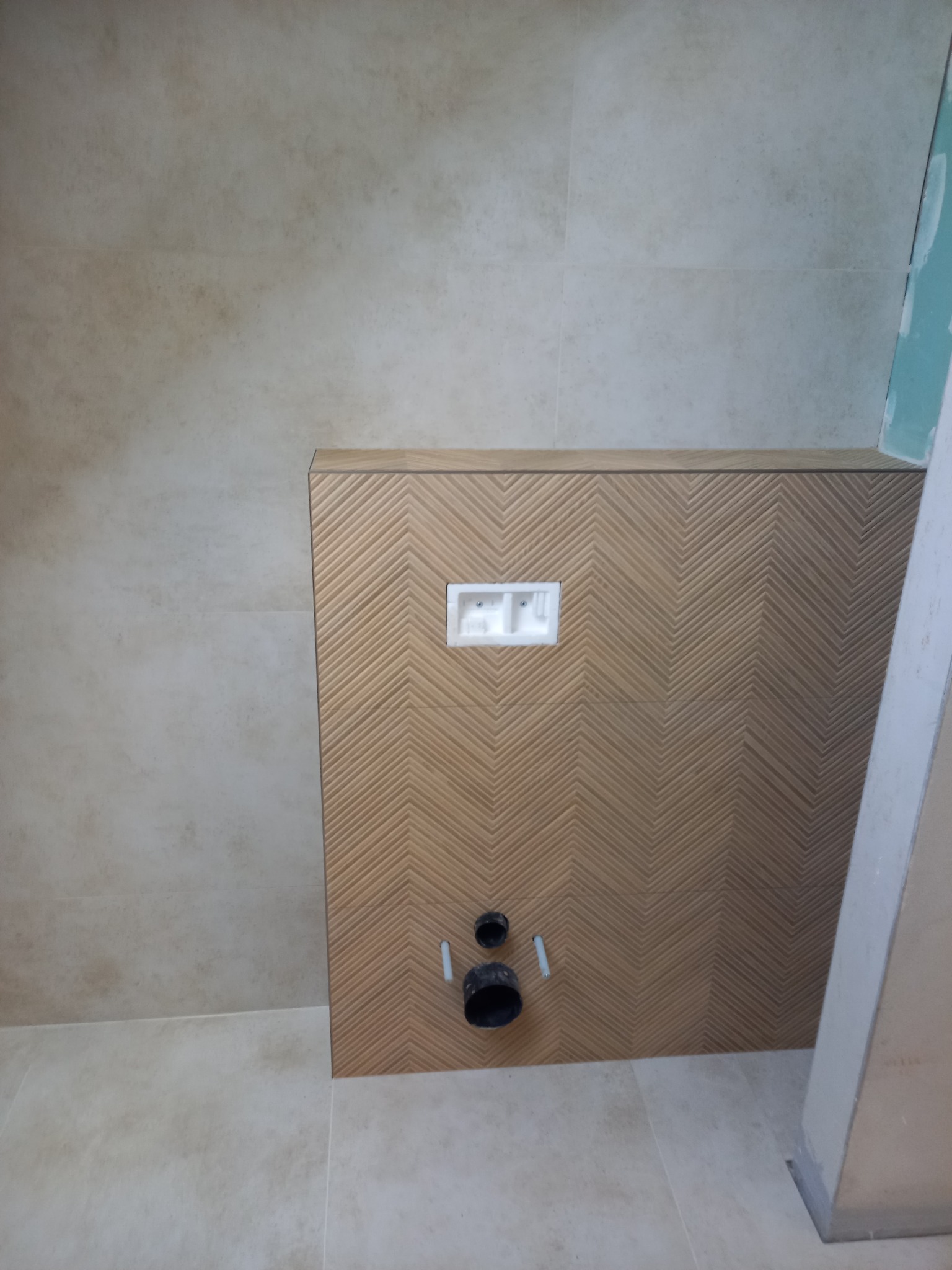 Wykończenie ściany w łazience z dekoracyjną okładziną w jodełkę, widoczne przyłącza do toalety podtynkowej i fragment płyty gipsowej. Neutralna kolorystyka.