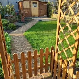 Ogród z drewnianą altaną i ścieżką z kostki, prowadzącą do domku narzędziowego. Starannie utrzymany trawnik i rabaty kwiatowe w słoneczny dzień.