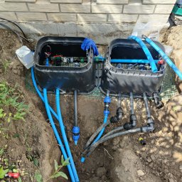 JDG Andriy Grytsenko - Dwa czarne zawory nawadniające w ziemi, połączone niebieskimi i czarnymi rurami, z widocznymi elementami instalacji i narzędziami. Częściowo odsłonięty system.