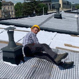 JDG Andriy Grytsenko - Pracownik w kasku siedzi na dachu pokrytym siatką i membraną, obok komin. W tle budynki miejskie i błękitne niebo. Widok z góry.