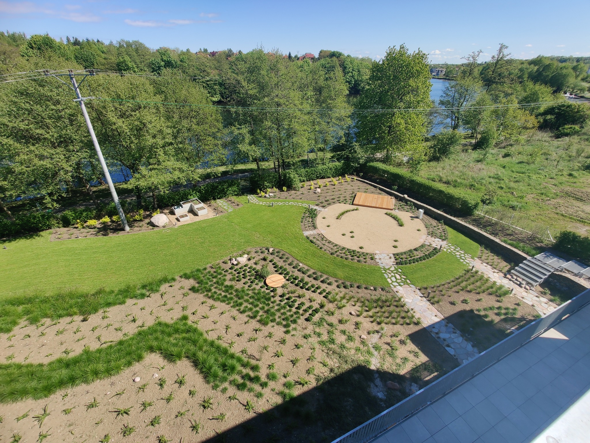 Widok z góry na starannie zaprojektowany ogród z okrągłym tarasem, ścieżkami z kamieni i różnorodną roślinnością, na tle zieleni i zbiornika wodnego.