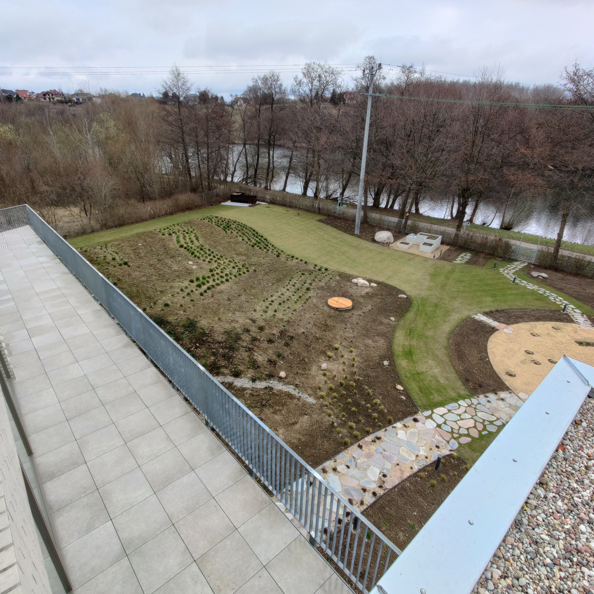 Widok z góry na nowo założony ogród w Tczewie: trawnik, ścieżki z kamienia, nasadzenia w rzędach i drzewa w tle. Elementy małej architektury.