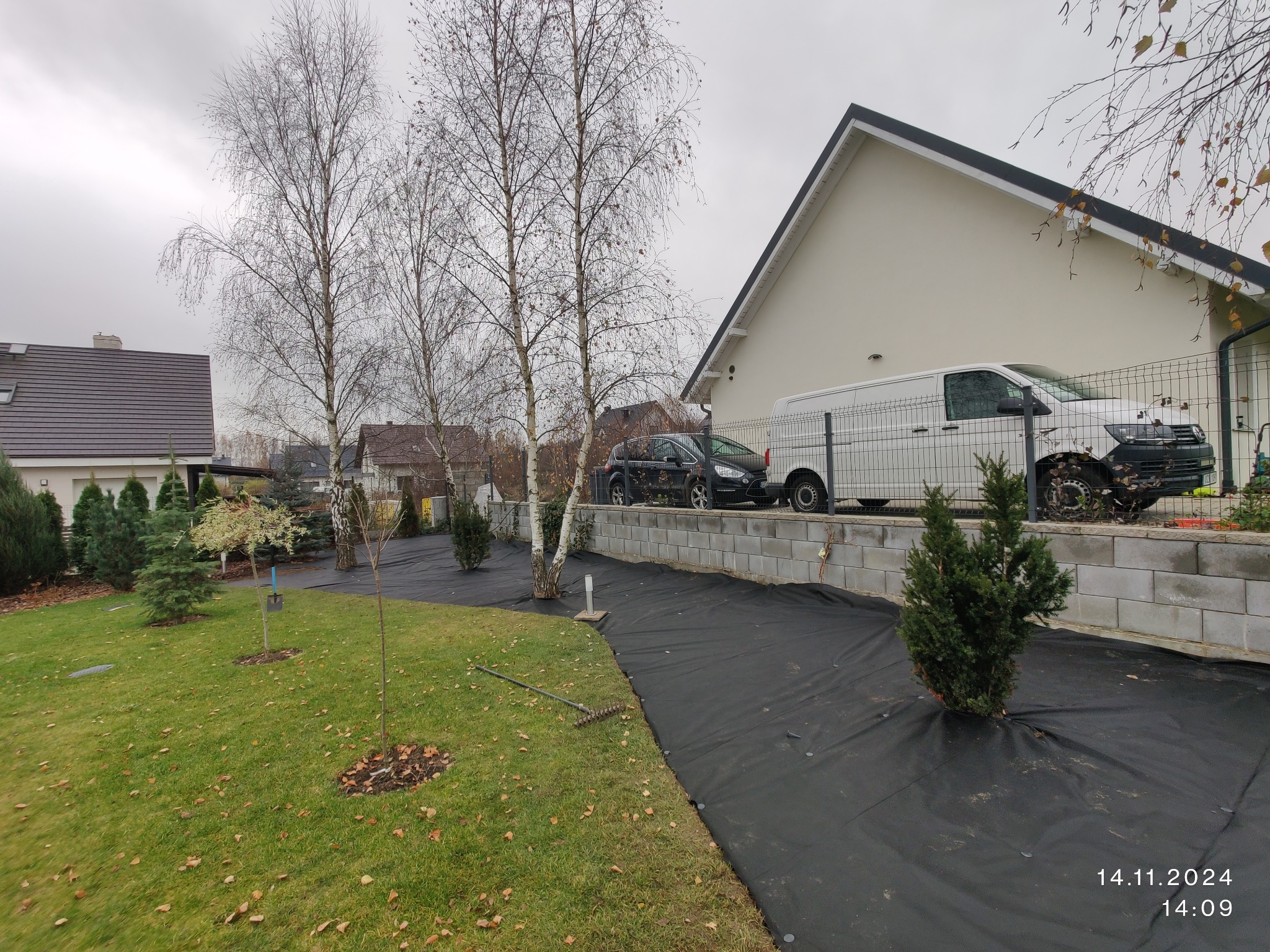 Przygotowanie terenu pod przyszły ogród: rozłożona czarna agrowłóknina, młode drzewka, trawnik z opadłymi liśćmi, budynek w tle i zaparkowany samochód.