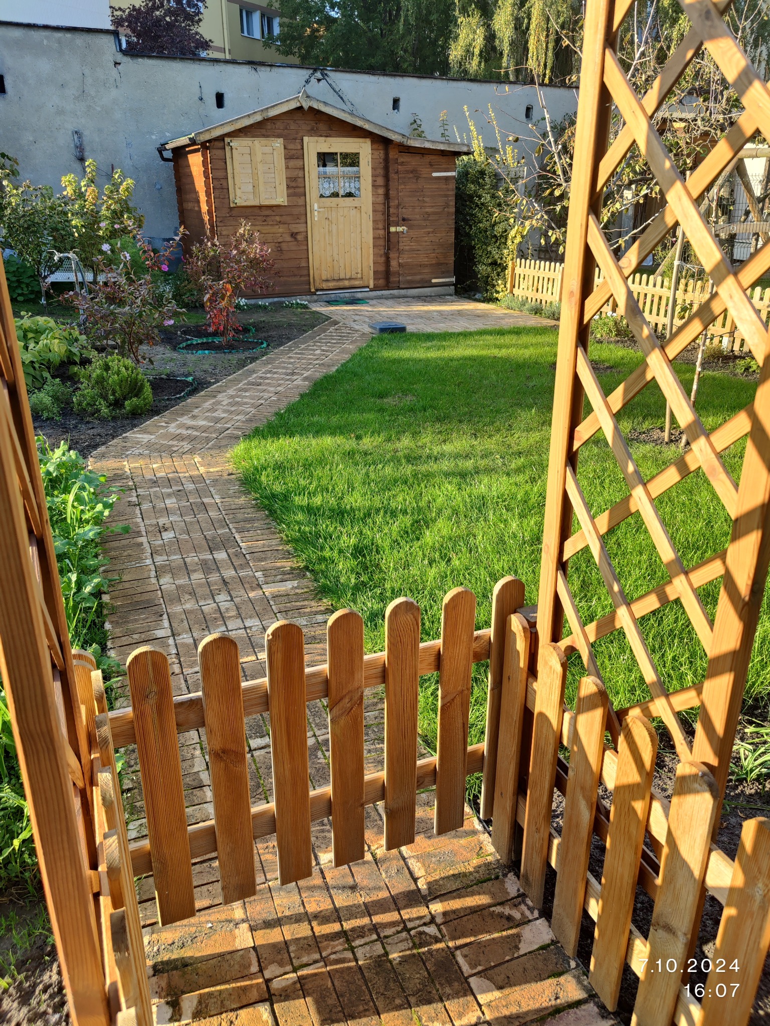 Ogród z drewnianą altaną i ścieżką z kostki, prowadzącą do domku narzędziowego. Starannie utrzymany trawnik i rabaty kwiatowe w słoneczny dzień.