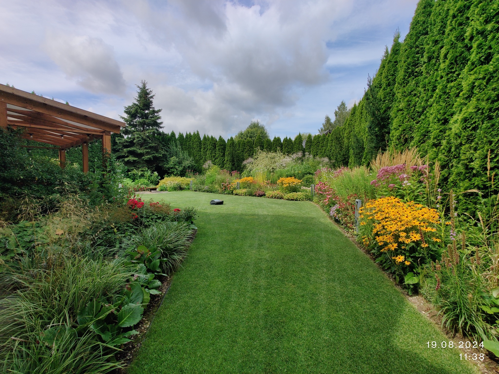 Zadbany ogród z równo przystrzyżonym trawnikiem, robotem koszącym, rabatami kwiatowymi i żywopłotem z tuj. Widok na drewnianą altanę i niebo z chmurami.