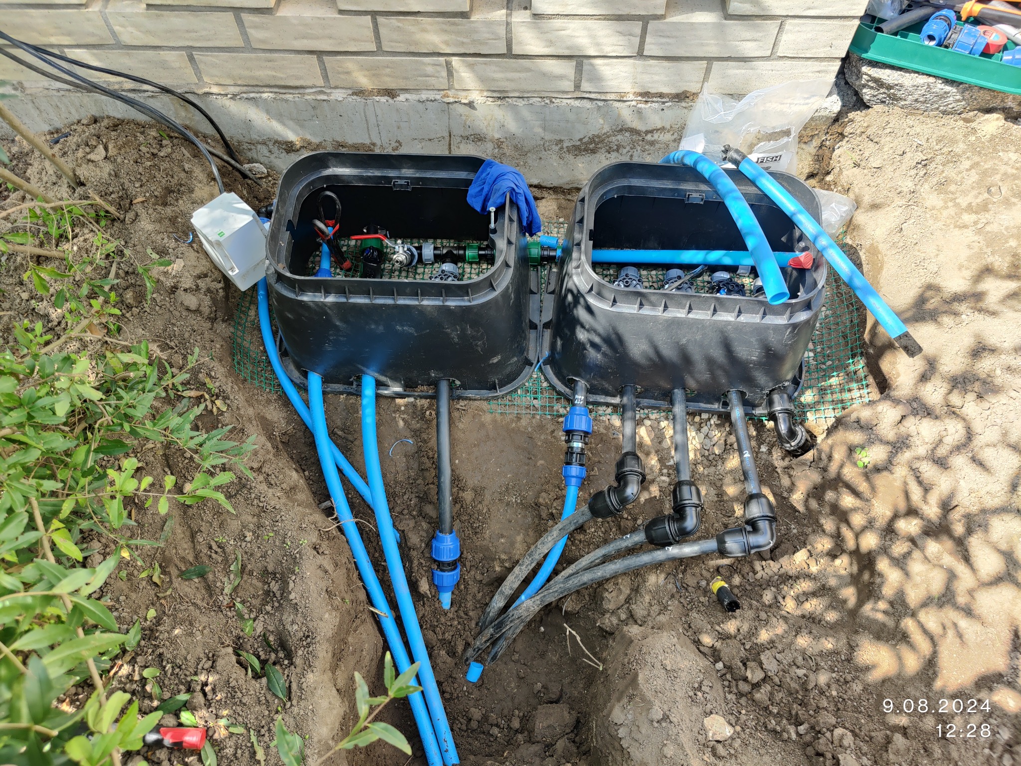 Dwa czarne zawory nawadniające w ziemi, połączone niebieskimi i czarnymi rurami, z widocznymi elementami instalacji i narzędziami. Częściowo odsłonięty system.
