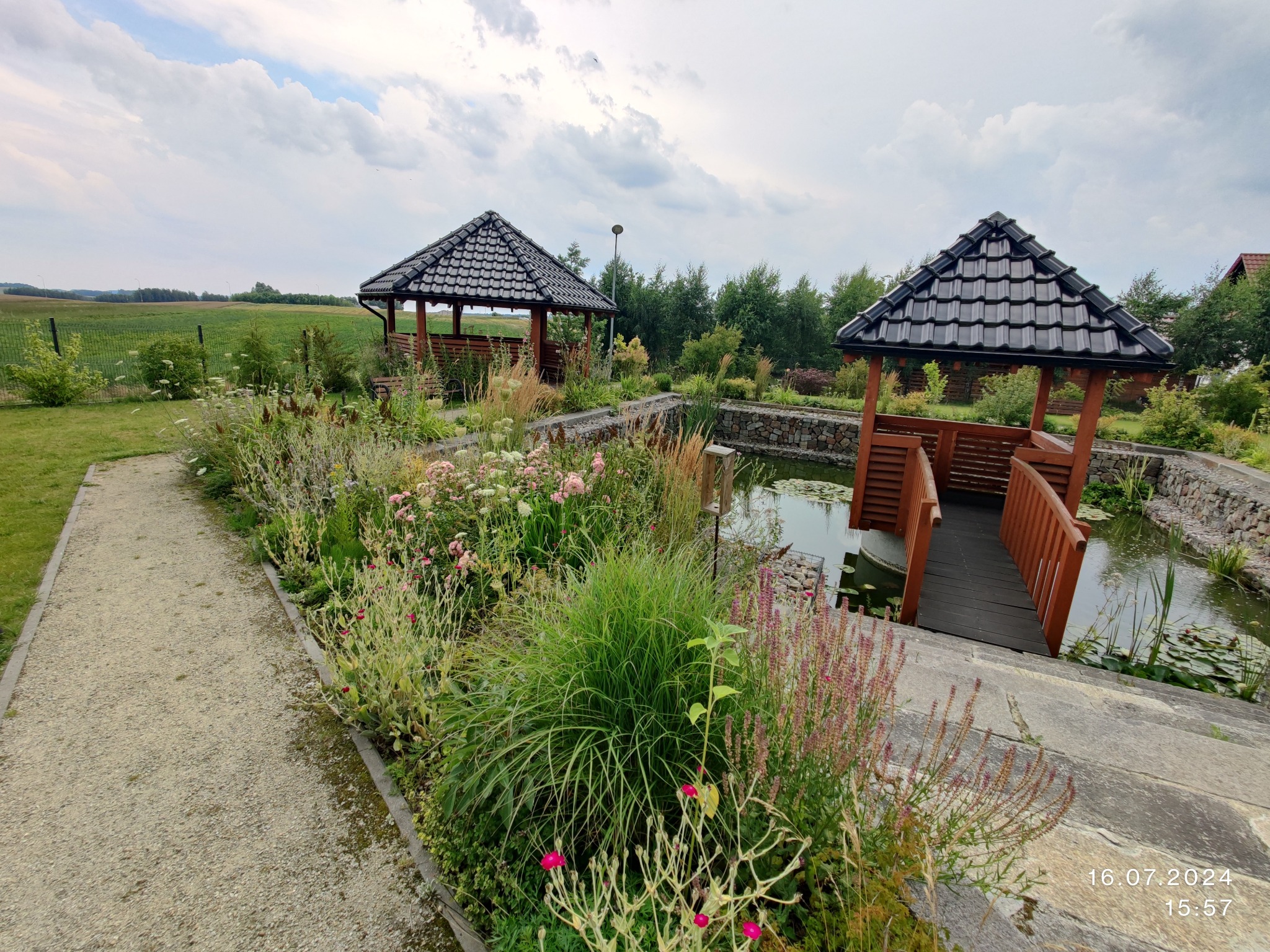 Ogród z dwoma altanami nad stawem, kamienne obrzeża, ścieżka żwirowa i kwiaty. Widok na zielone pola w tle pod pochmurnym niebem.