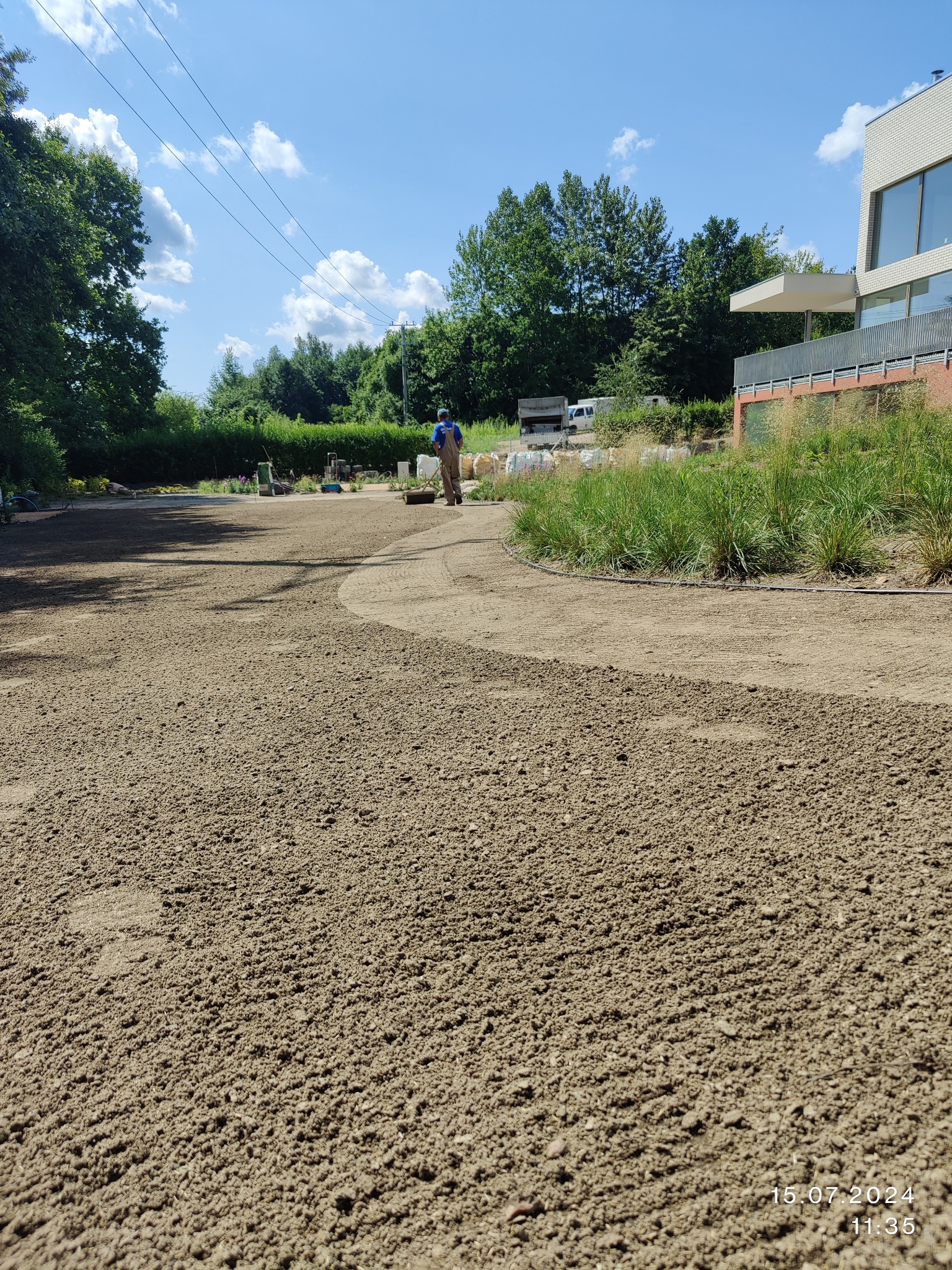 Przygotowany teren pod trawnik z wytyczonymi ścieżkami. Pracownik wyrównuje podłoże walcem na tle zieleni i nowoczesnej architektury w słoneczny dzień.