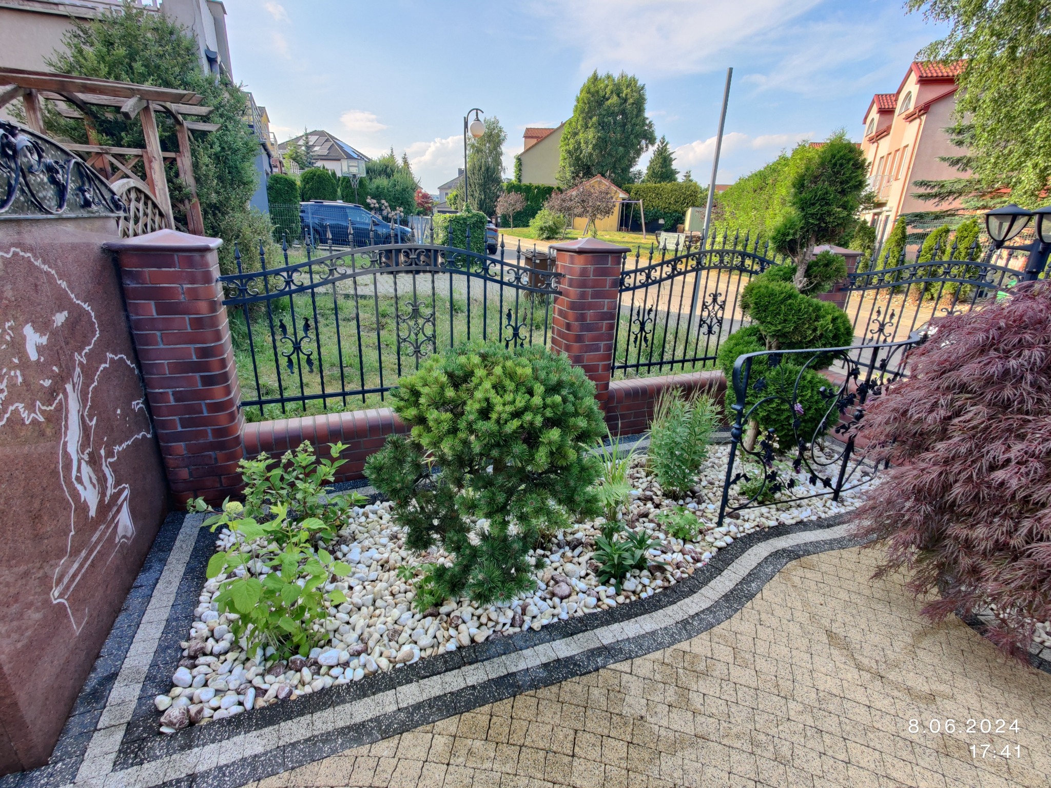 Kaskadowy ogródek skalny z ozdobnymi roślinami i białymi kamykami, obramowany kostką brukową. W tle metalowe ogrodzenie z ceglanymi słupkami.