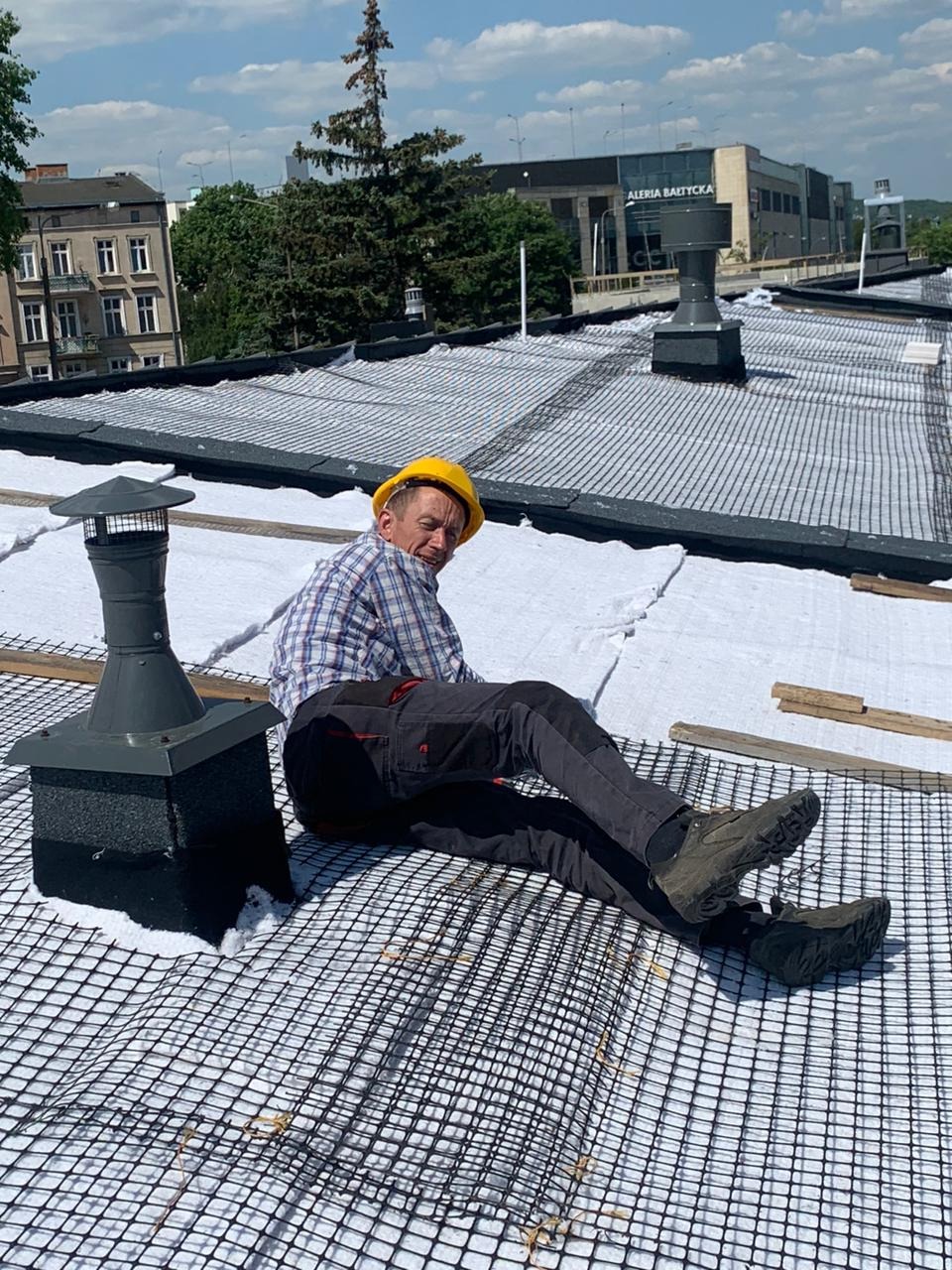 Pracownik w kasku siedzi na dachu pokrytym siatką i membraną, obok komin. W tle budynki miejskie i błękitne niebo. Widok z góry.