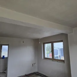 Wnętrze po tynkowaniu: gładkie, jasnoszare ściany, okno z widokiem na zewnątrz, widoczne instalacje w podłodze, drabina oparta o ścianę. Gotowe do dalszych prac wykończeniowych.