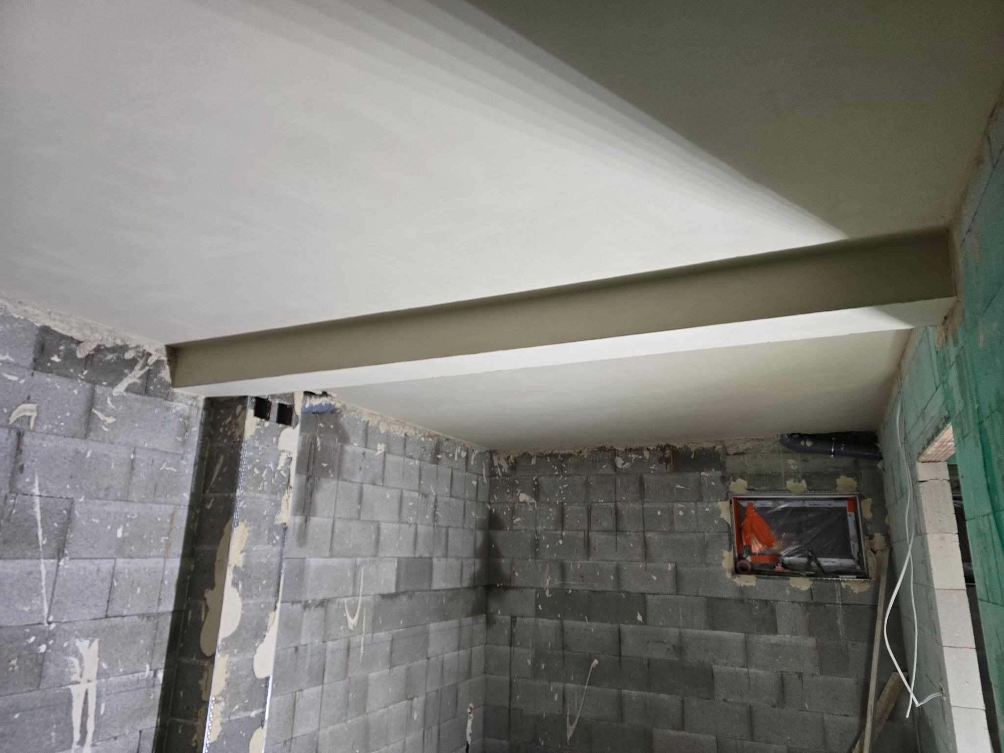 Wnętrze surowego pomieszczenia z wygładzonym, jasnym tynkiem na suficie i fragmentami ścian. Widoczne bloczki betonowe i stalowy element konstrukcyjny pod sufitem.