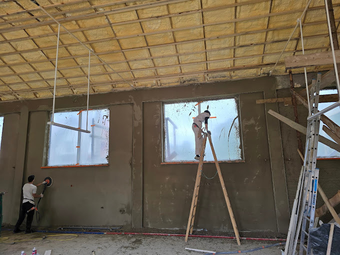 Prace tynkarskie wewnątrz budynku: Dwóch mężczyzn przy oknach, jeden na drabinie, drugi szlifuje ścianę. Widoczna surowa struktura sufitu i plastikowe zabezpieczenia okien.