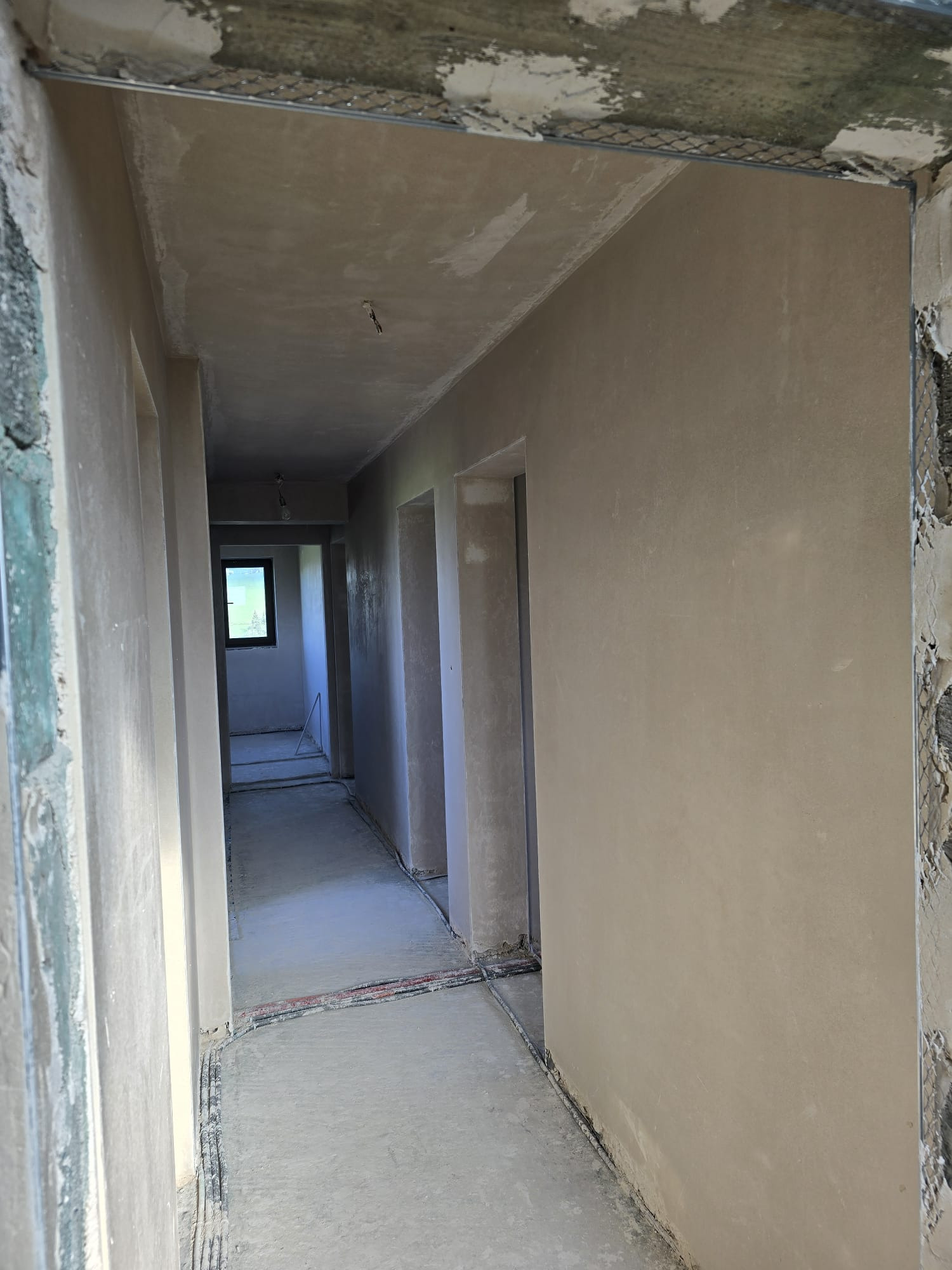 Wykończony korytarz z gładkimi, jasnymi tynkami maszynowymi na ścianach. Widoczne kable wzdłuż podłogi i ościeżnice drzwiowe. W głębi okno z widokiem na zieleń.