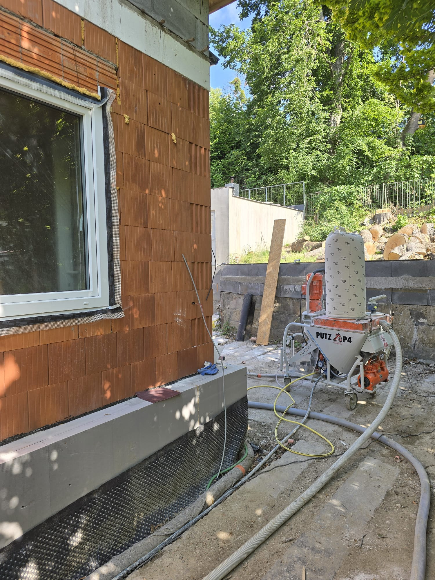 Praca z agregatem tynkarskim Putz P4 na budowie domu z cegły. Widoczna ściana z oknem w trakcie przygotowań do tynkowania.