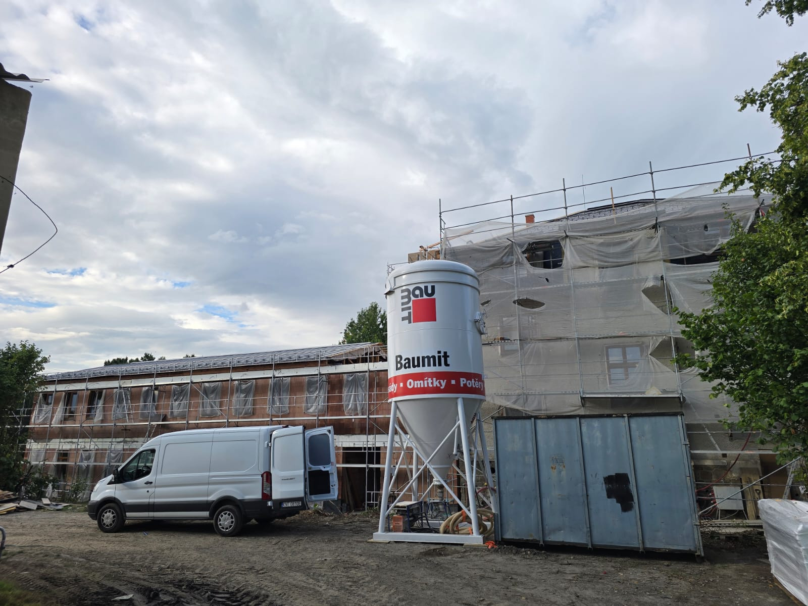 Prace tynkarskie na budynku w rusztowaniach, widoczny silos Baumi z otwartym busem dostawczym. Zabezpieczenie folią. Ujęcie z dołu, pochmurne niebo.